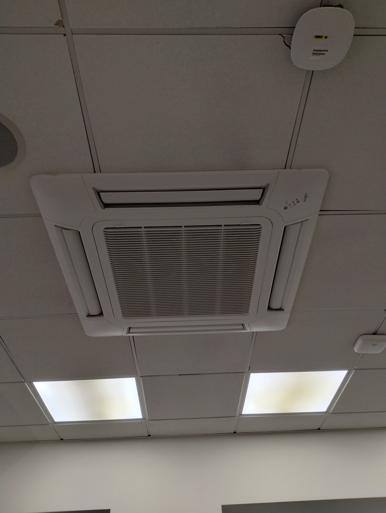 Kasetonowa klimatyzacja sufitowa marki Fujitsu w biurze, widok z dołu. Biała obudowa, kratka wentylacyjna. Widoczne panele sufitowe i oświetlenie.