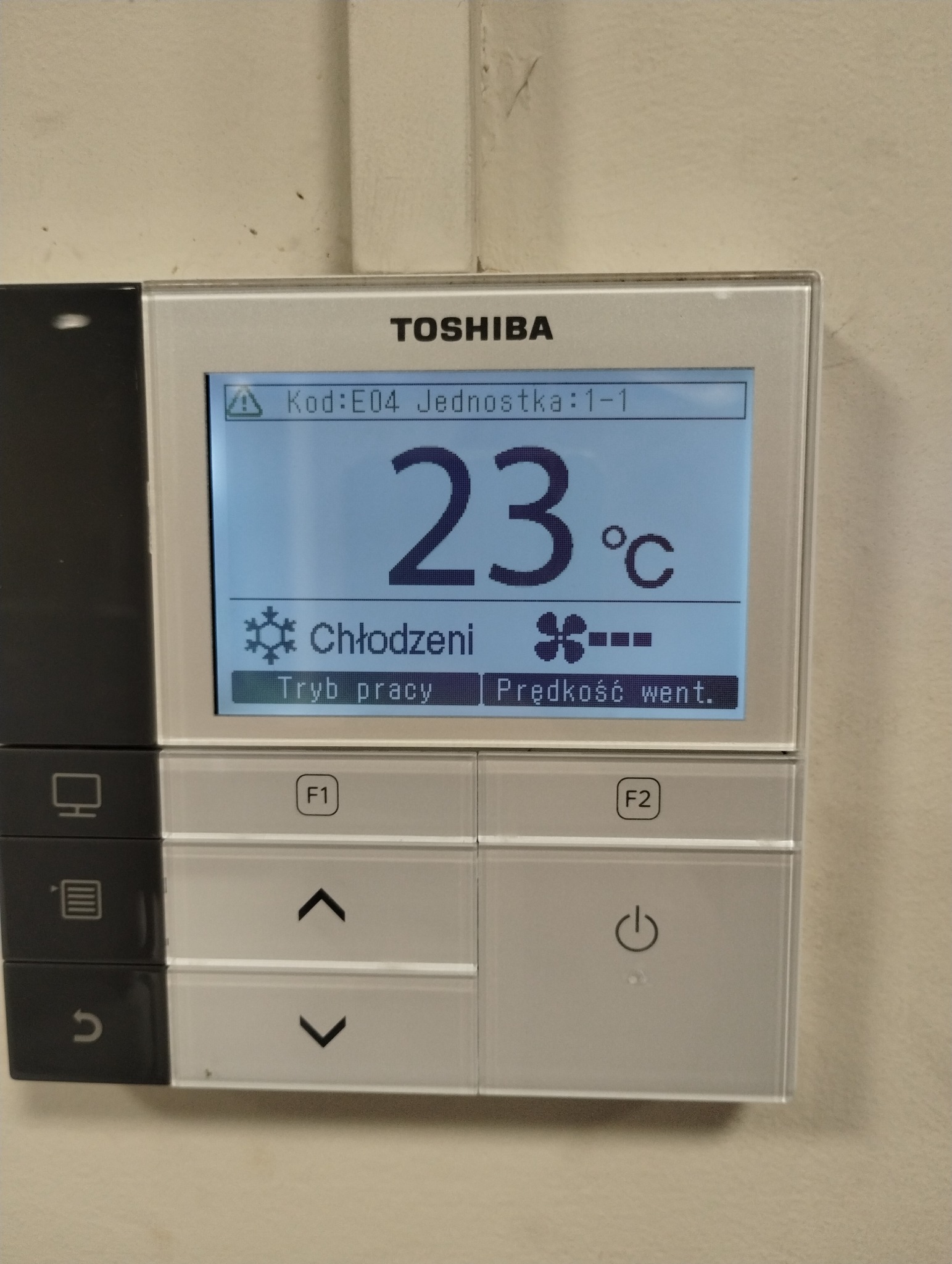 Panel sterowania klimatyzacji Toshiba z wyświetlaną temperaturą 23°C, trybem chłodzenia i regulacją prędkości wentylatora. Widoczne przyciski funkcyjne F1 i F2.