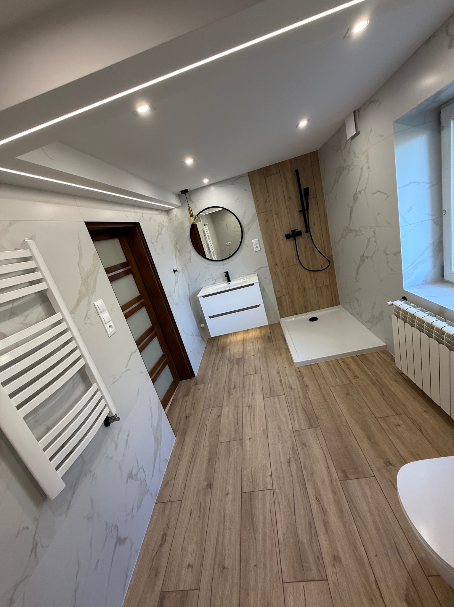 Nowoczesna łazienka z drewnopodobną podłogą, marmurowymi płytkami, kabiną prysznicową typu walk-in i podświetlanym lustrem. Grzejnik drabinkowy i oświetlenie LED.