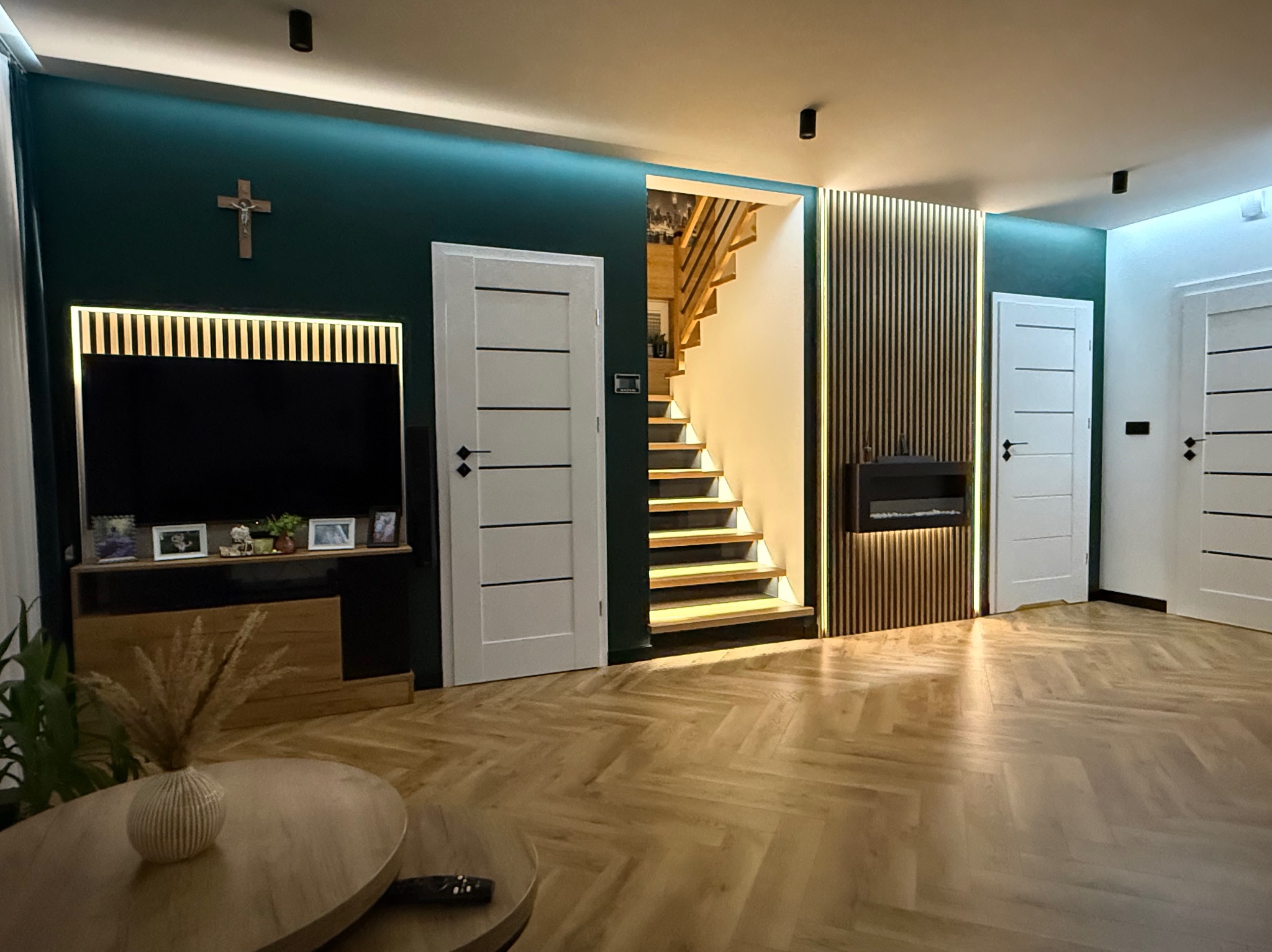 Salon z parkietem ułożonym w jodełkę, zielona ściana z krzyżem, dwoje białych drzwi z czarnymi klamkami, schody z drewnianymi stopniami i podświetleniem LED.