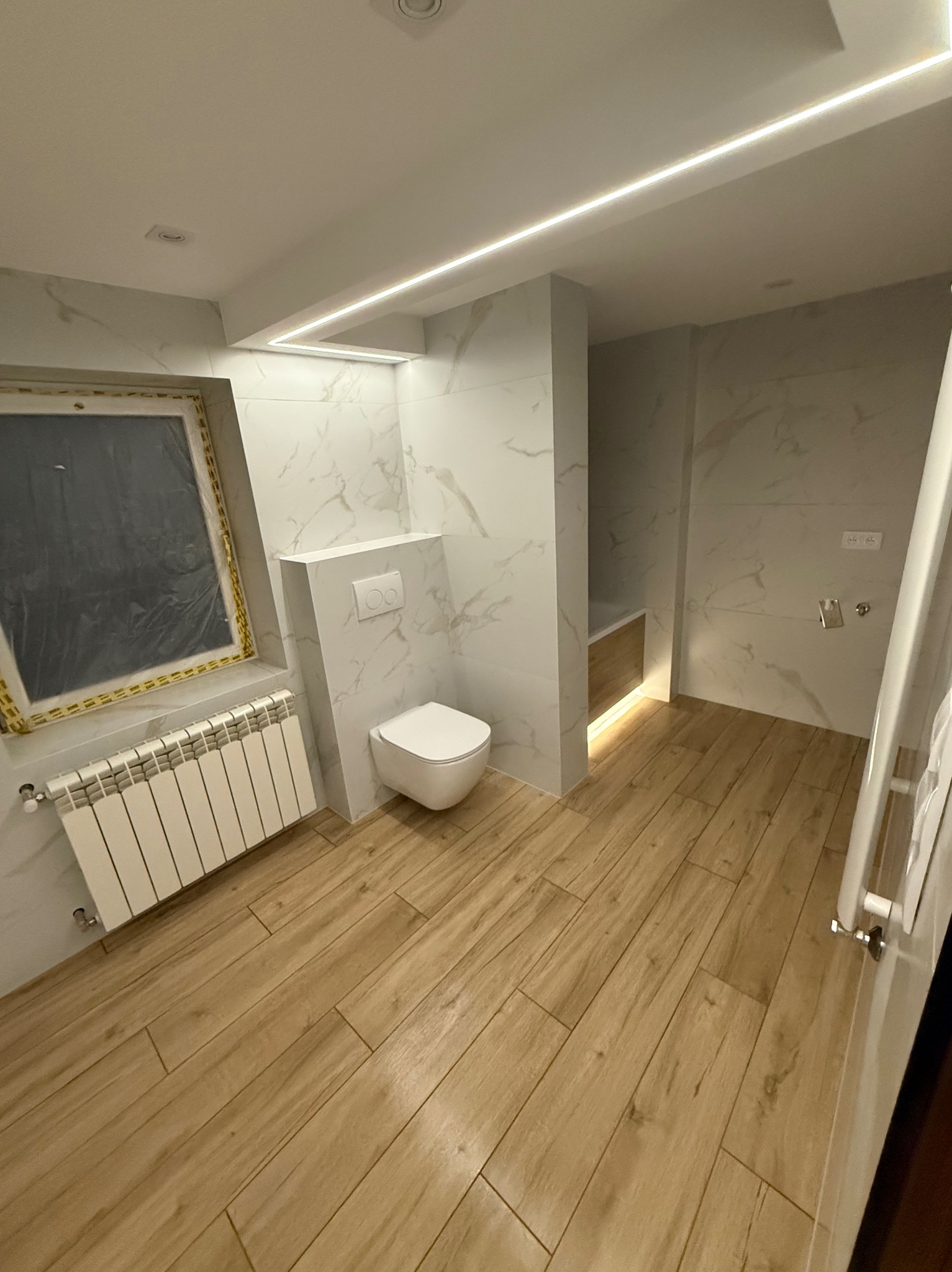 Nowoczesna łazienka po remoncie: białe marmurowe płytki, drewnopodobna podłoga, podświetlenie LED, wisząca toaleta i grzejnik pod oknem z folią ochronną.