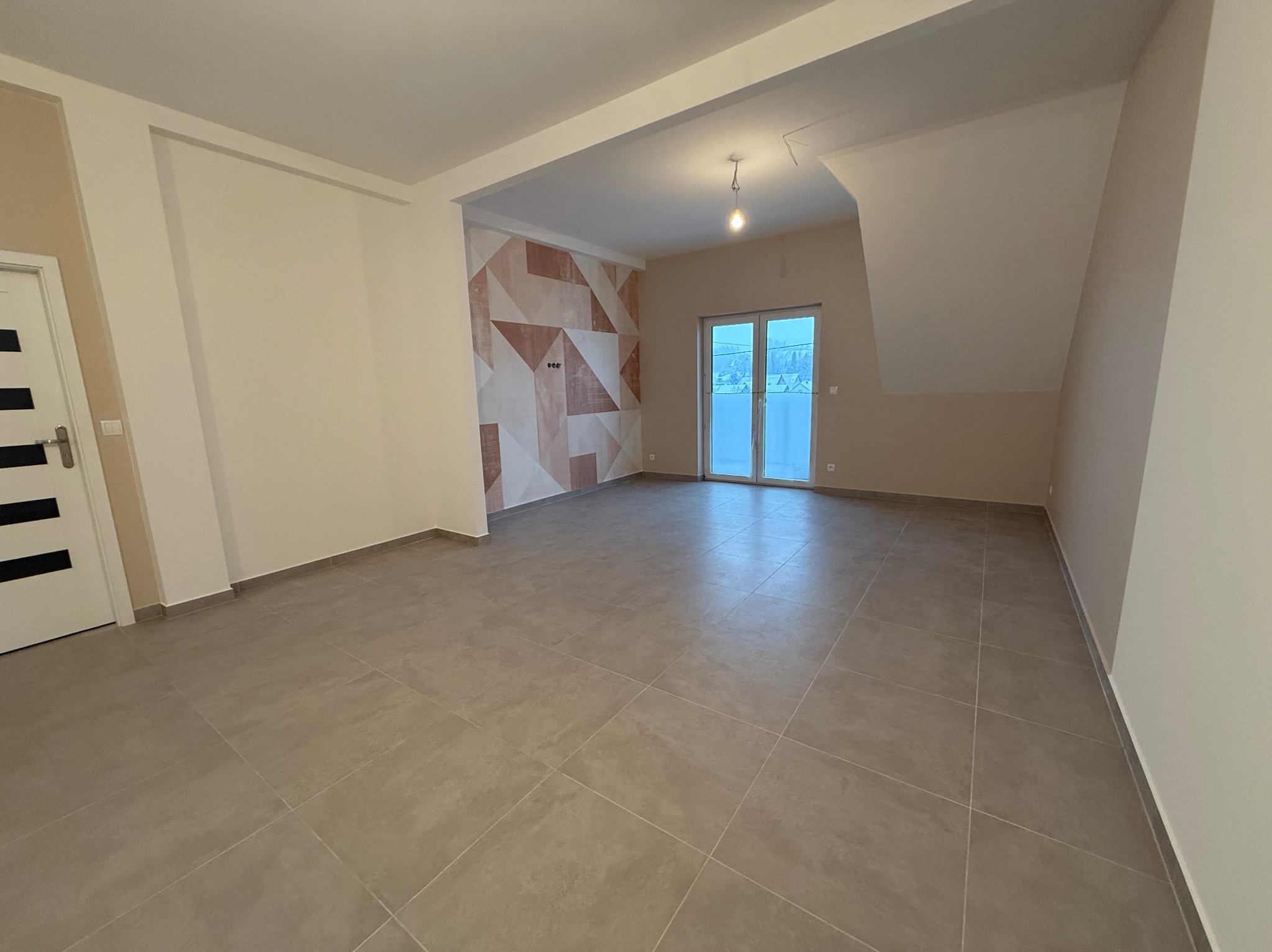 Wykończone wnętrze: beżowe płytki podłogowe, ściana z geometrycznym wzorem w odcieniach brązu i beżu, białe drzwi z czarnymi akcentami, okno balkonowe.