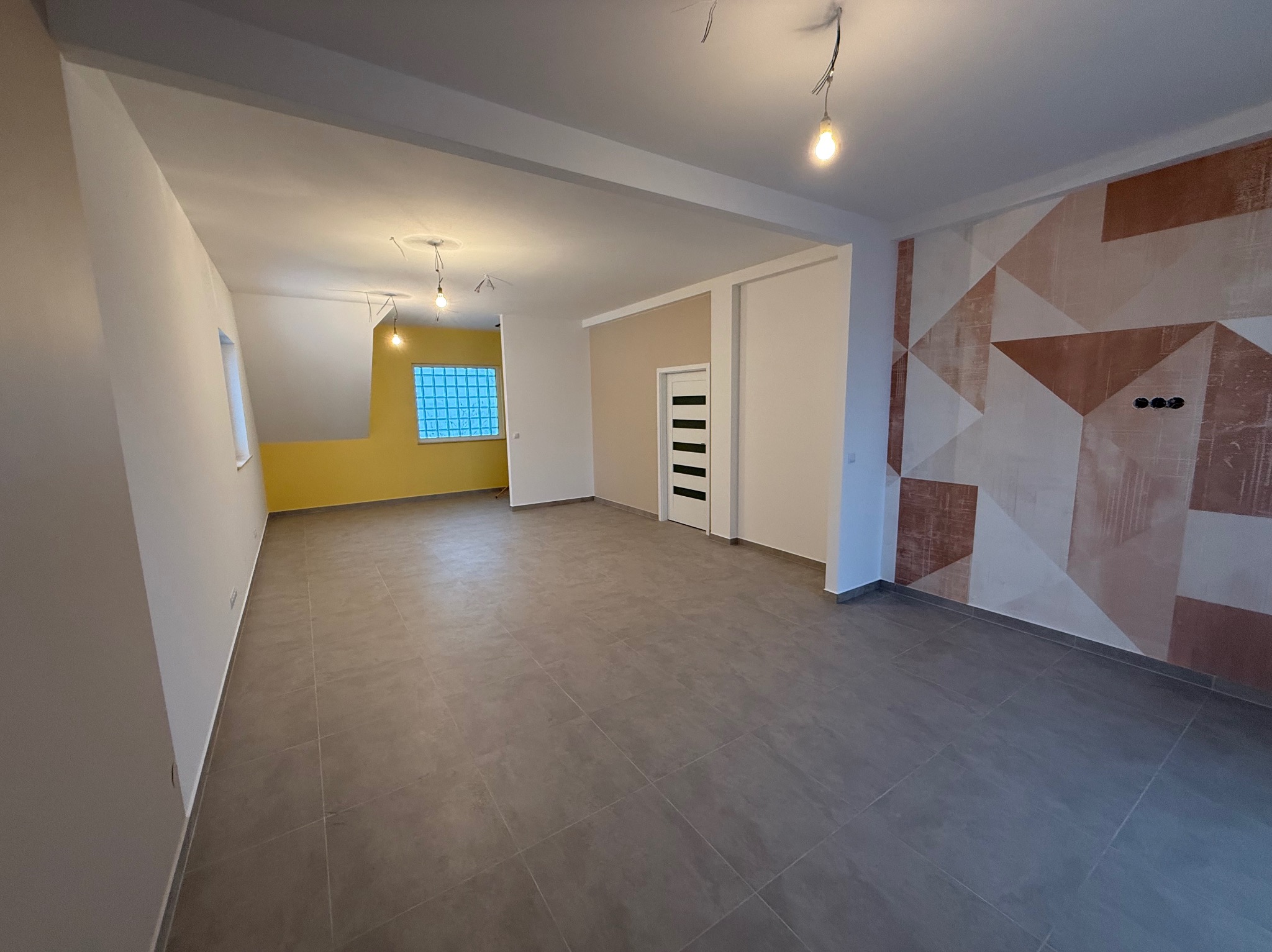 Wyremontowane, przestronne wnętrze z szarą podłogą, geometryczną ścianą akcentującą i żółtym pomieszczeniem w tle. Widoczne lampy wiszące i białe drzwi.