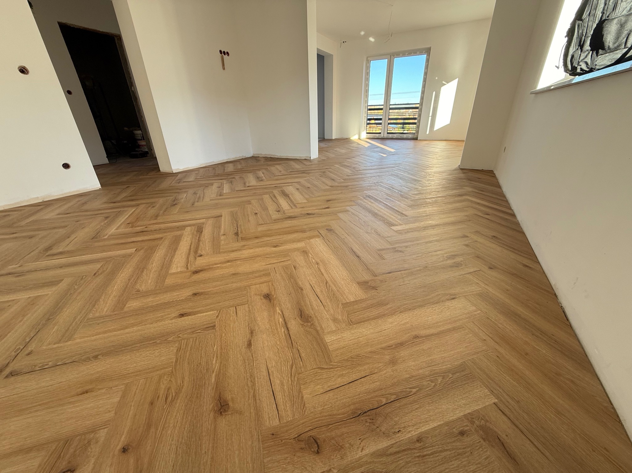 Jasny parkiet ułożony we wzór jodełki francuskiej w przestronnym, słonecznym wnętrzu. Widoczne surowe ściany i przygotowane otwory na gniazdka elektryczne.