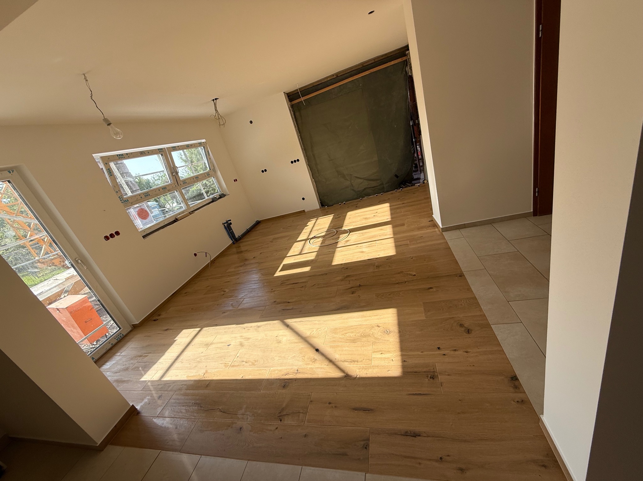 Jasne, przestronne wnętrze z ułożonym parkietem w odcieniach beżu i brązu. Widoczne okno z widokiem na zewnątrz i przygotowane punkty elektryczne na ścianach.