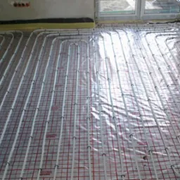 Rozłożona instalacja ogrzewania podłogowego na siatce z tworzywa, widoczne rury w kształcie pętli przed zalaniem wylewką, okno w tle.