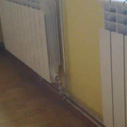 Dwa białe grzejniki panelowe zamontowane przy ścianie z widocznymi rurami doprowadzającymi wodę, ujęcie z niskiej perspektywy.