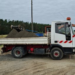 AGBETKOM AGNIESZKA KOMOROWSKA - Ciężarówka z wywrotką wypełniona ziemią na placu budowy, z widocznym sprzętem do załadunku i lasem w tle, pod pochmurnym niebem.