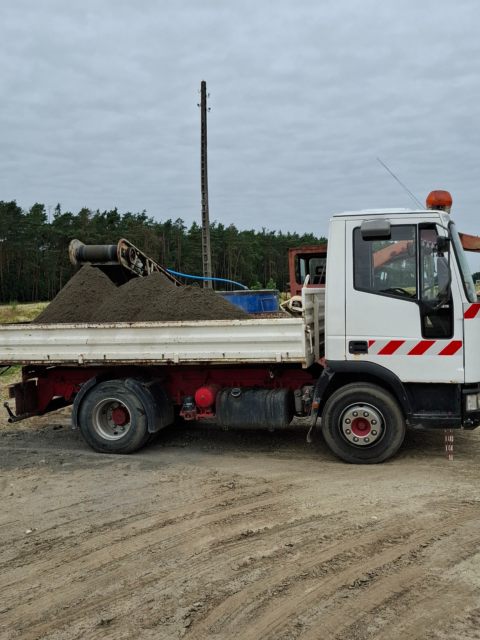 Ciężarówka z wywrotką wypełniona ziemią na placu budowy, z widocznym sprzętem do załadunku i lasem w tle, pod pochmurnym niebem.