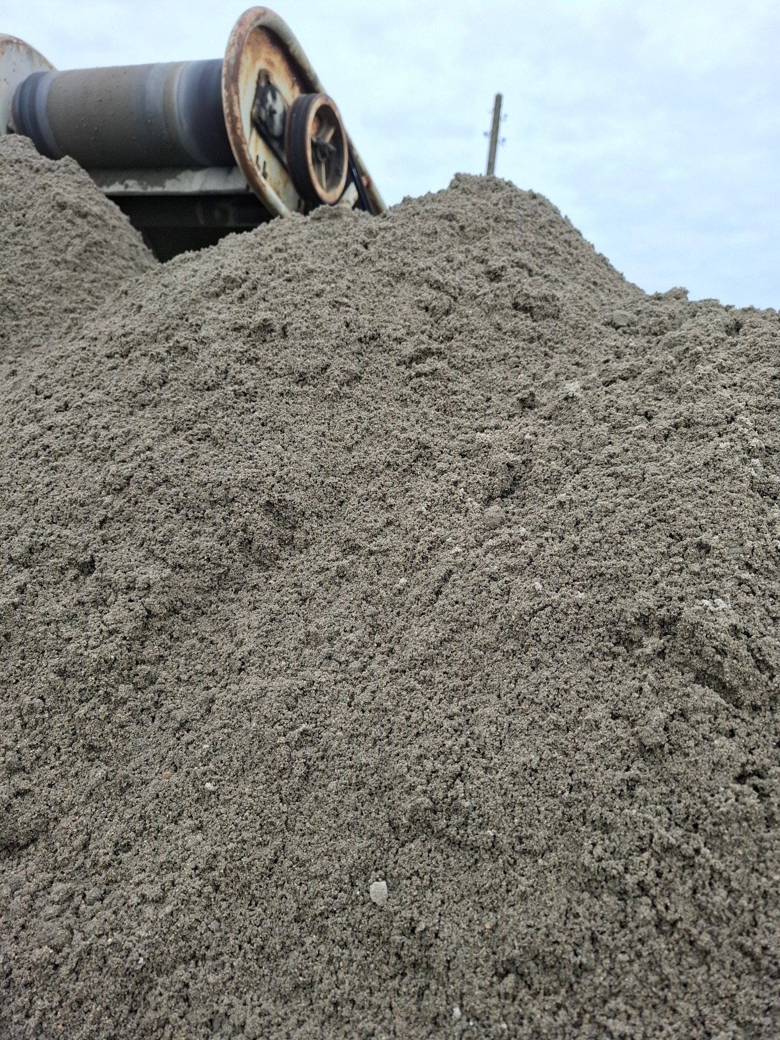 Góra piasku pod taśmociągiem z widocznymi elementami konstrukcyjnymi i zardzewiałymi detalami, na tle pochmurnego nieba. Materiał sypki w zbliżeniu.