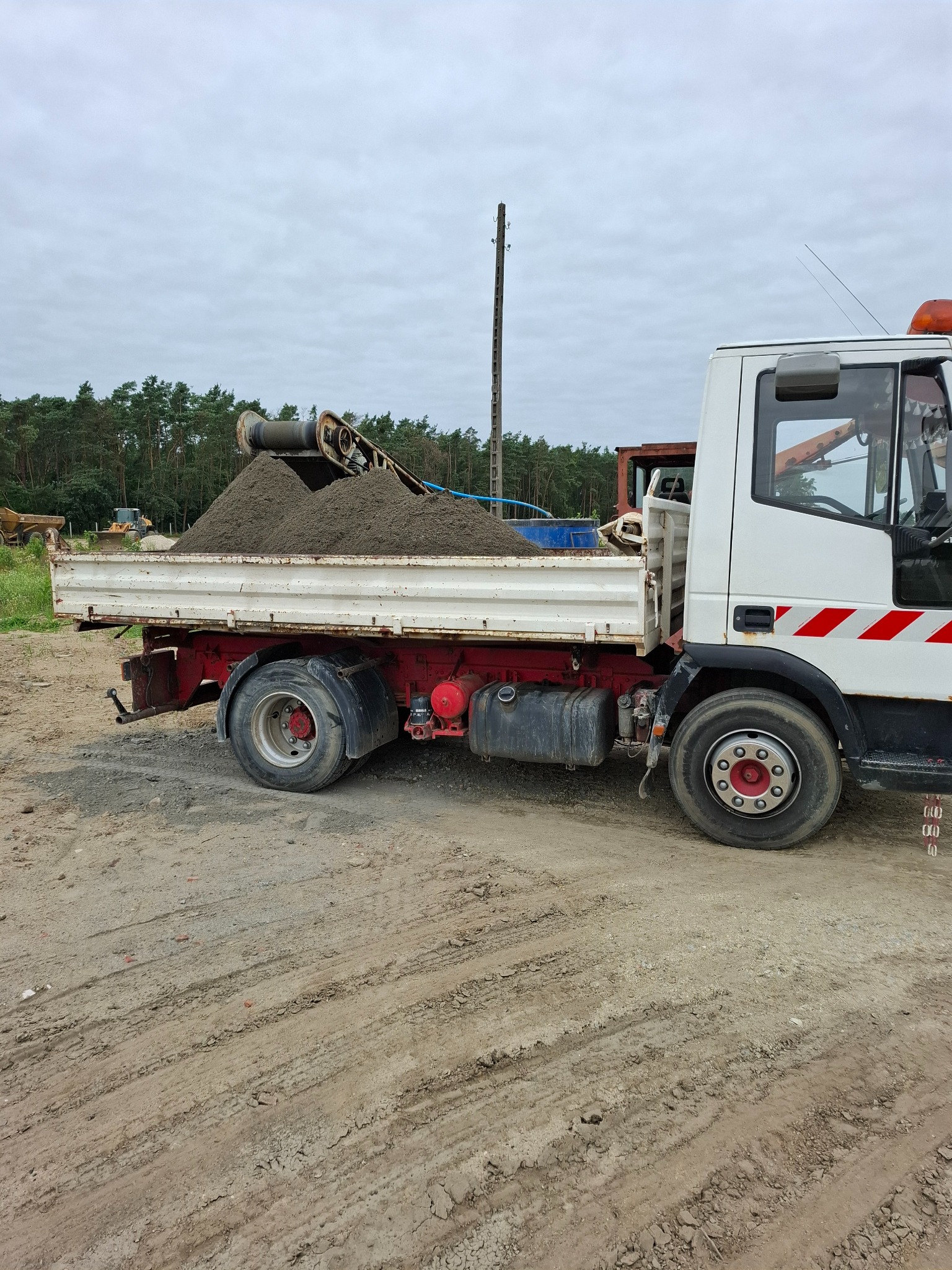 Ciężarówka z wywrotką wypełniona piaskiem na placu budowy, z lasem w tle i słupem energetycznym po lewej stronie. Widoczne ślady opon na ziemi.