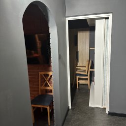 Ludwik usługi remontowe-elektryczne - Szare wnętrze z ceglaną ścianą widoczną przez łuk. Krzesło z ciemnym siedziskiem stoi przy ścianie. W tle widoczne kolejne krzesła i drzwi.