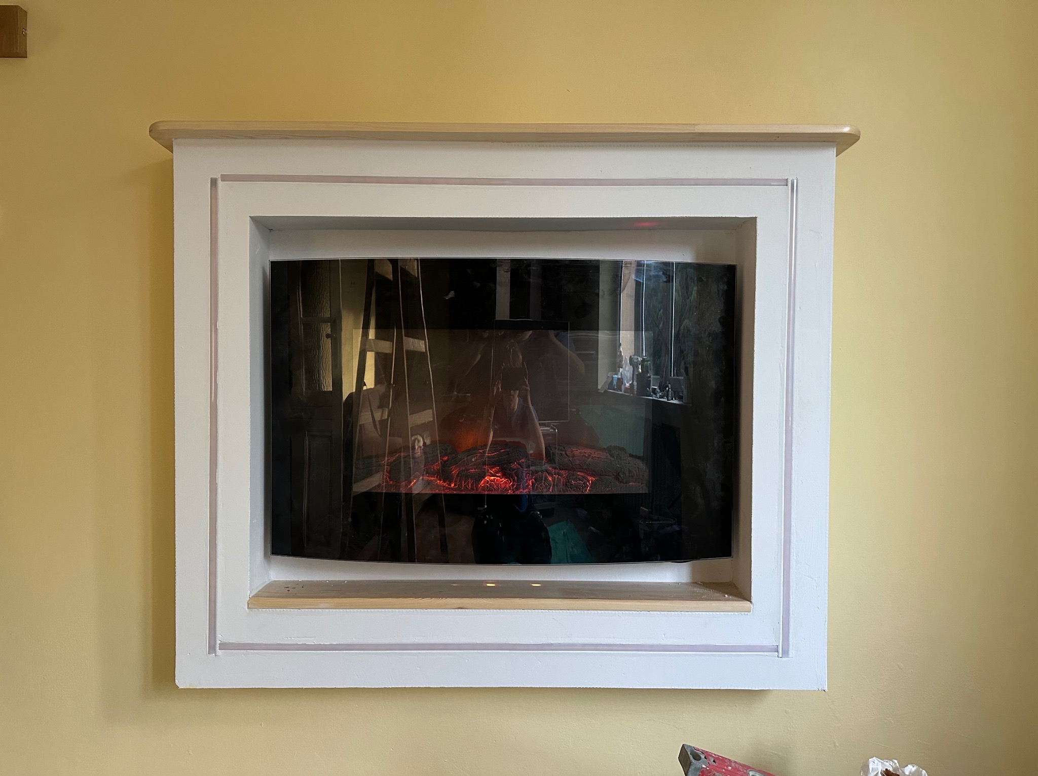 Biała obudowa kominka elektrycznego na żółtej ścianie. Widoczne niedomalowane elementy i surowe drewno. Detal wykończenia wnętrza.