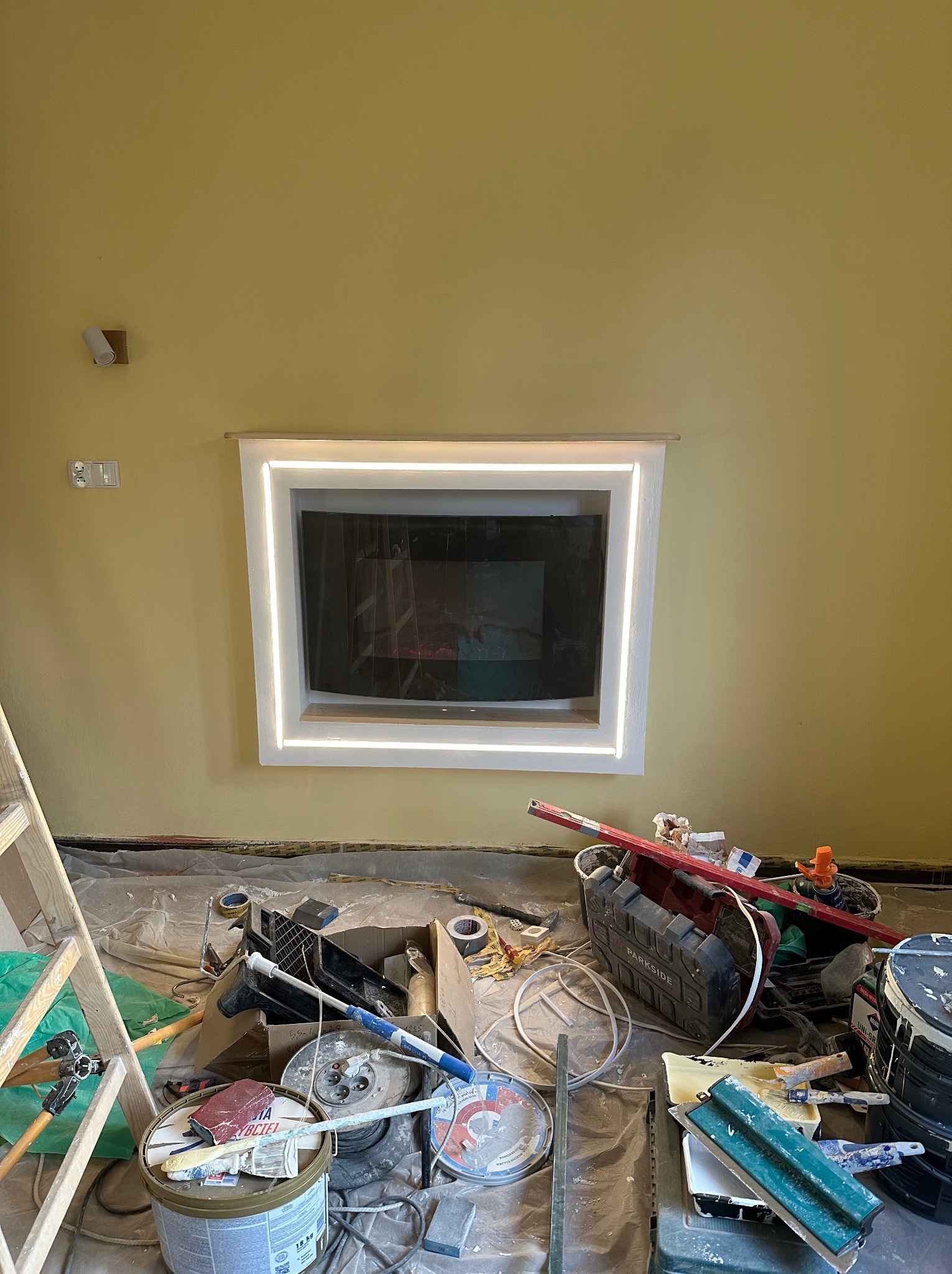 Ściana w trakcie prac wykończeniowych z wnęką oświetloną LED, narzędzia malarskie i drabina w pomieszczeniu o żółtych ścianach. Widok na wnętrze wnęki.