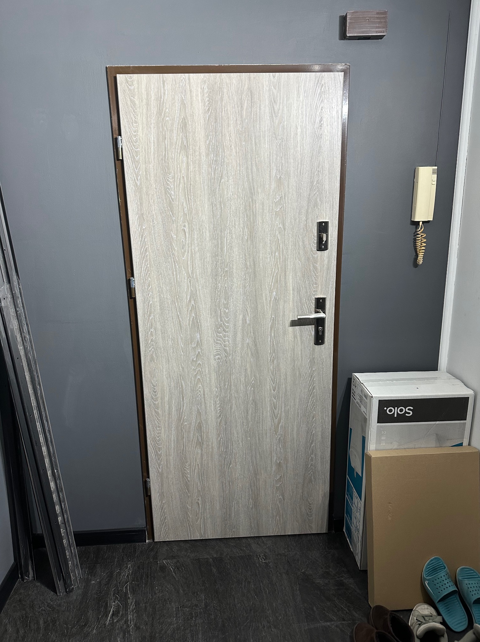 Zamknięte, jasne drzwi z drewnopodobnym wzorem, brązowa ościeżnica, szara ściana, domofon, kartony i listwy przypodłogowe w rogu. Widok na korytarz.