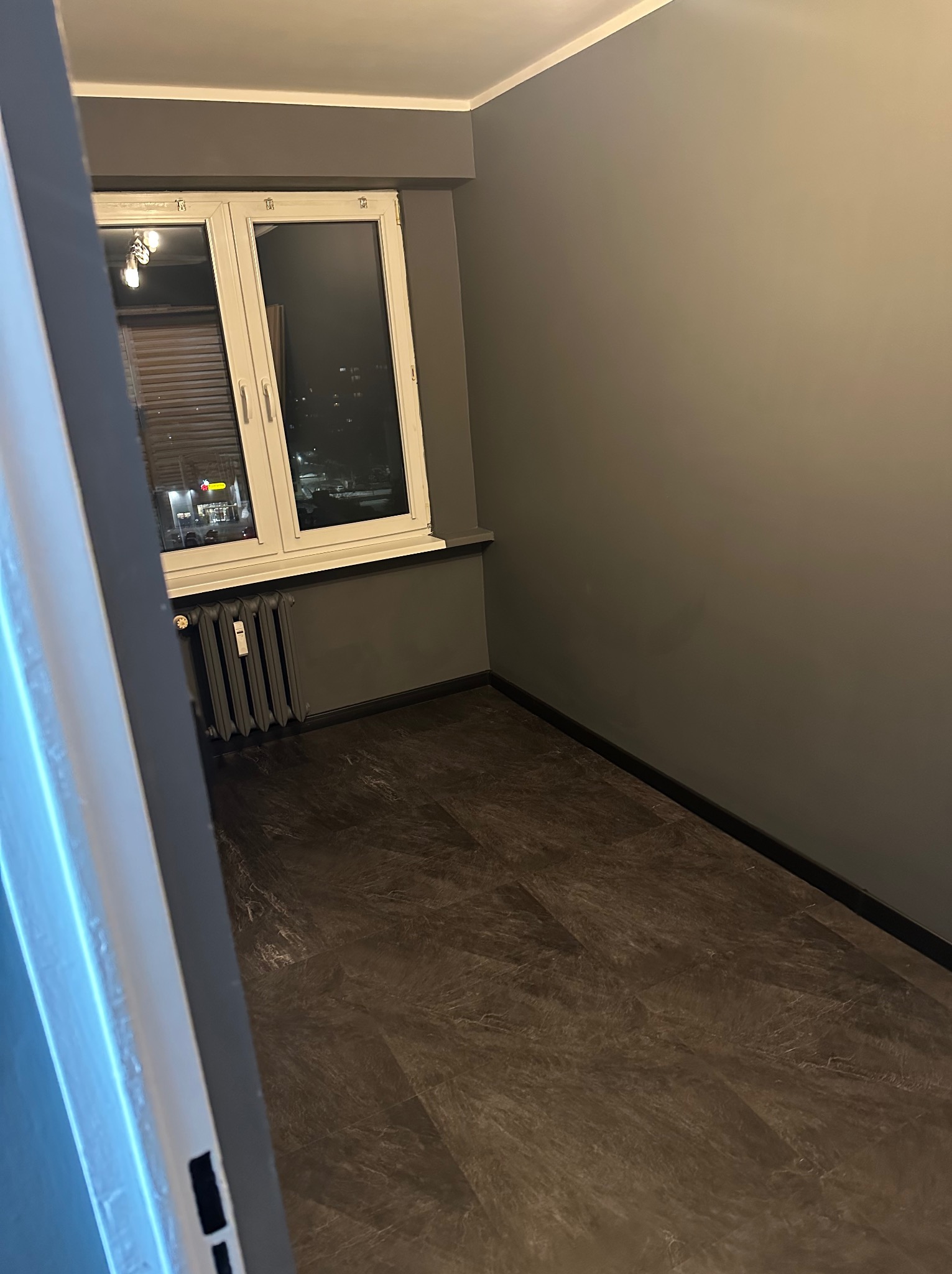 Pokój po remoncie w Kaliszu: ciemne panele podłogowe ułożone w jodełkę, szare ściany, białe okno z widokiem na nocne miasto. Grzejnik pomalowany na szaro.