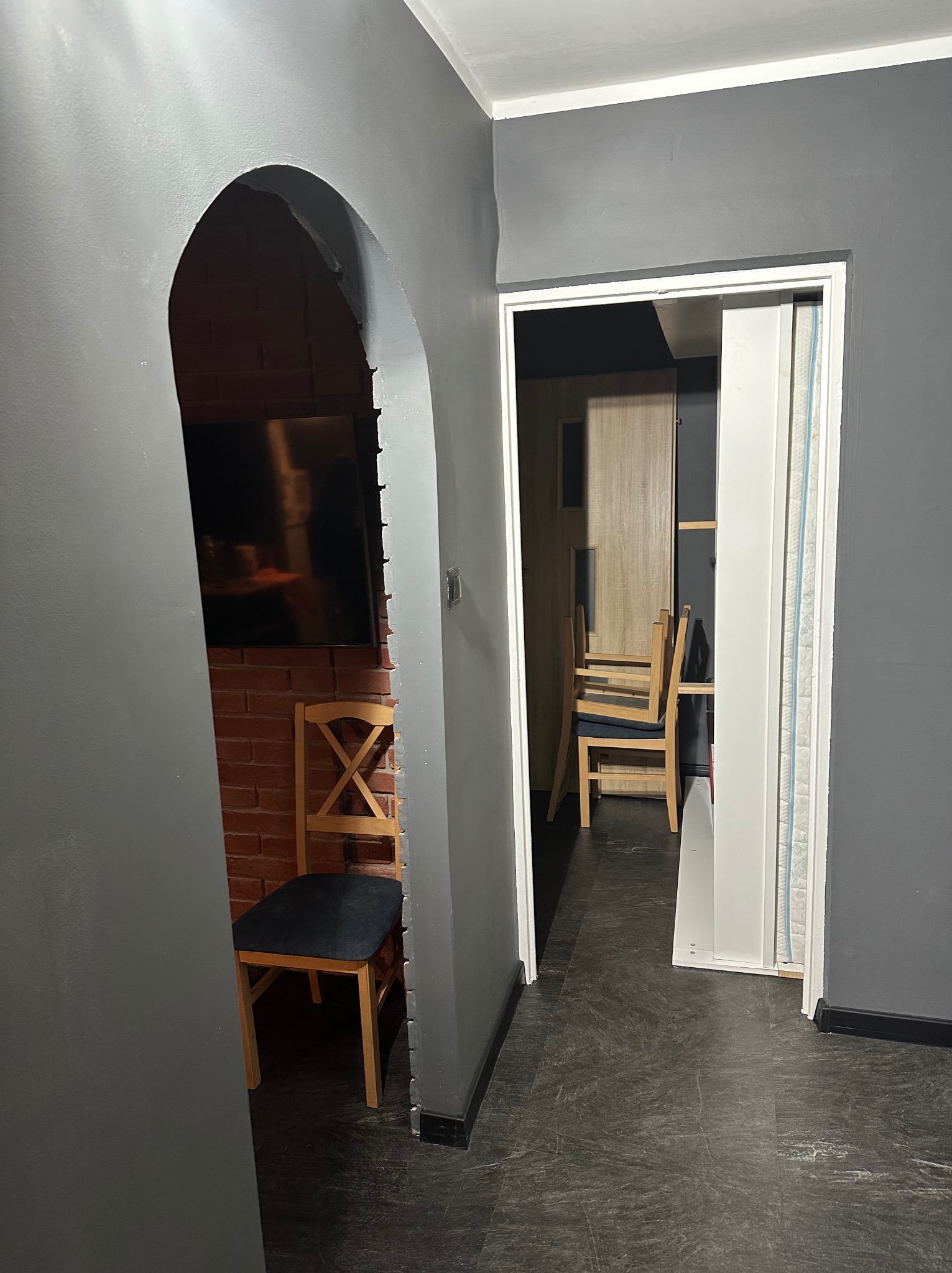 Szare wnętrze z ceglaną ścianą widoczną przez łuk. Krzesło z ciemnym siedziskiem stoi przy ścianie. W tle widoczne kolejne krzesła i drzwi.
