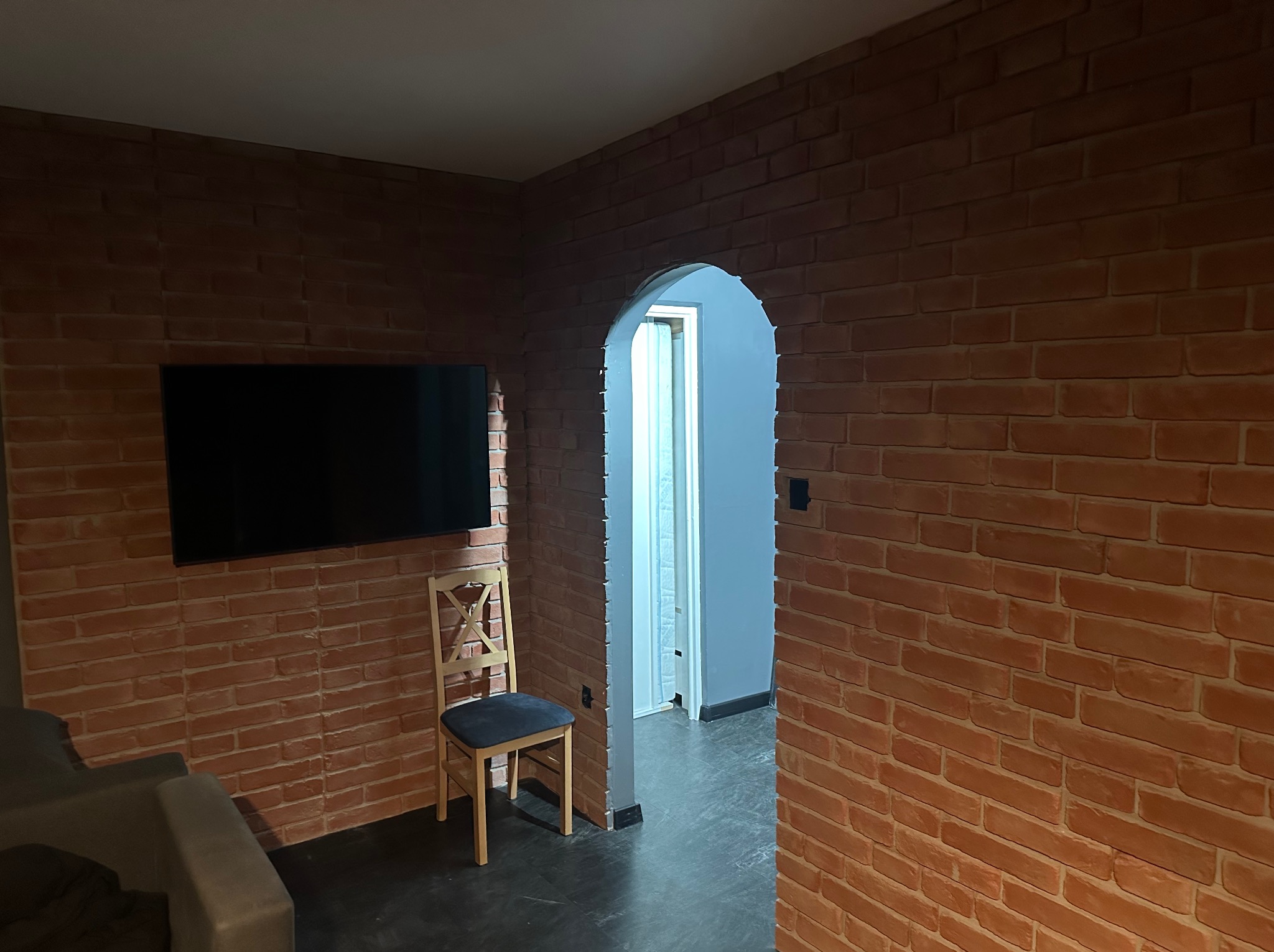 Ceglana ściana w salonie z telewizorem i krzesłem, widok na łukowate przejście do innego pomieszczenia. Dekoracyjne wykończenie wnętrza w stylu loft.