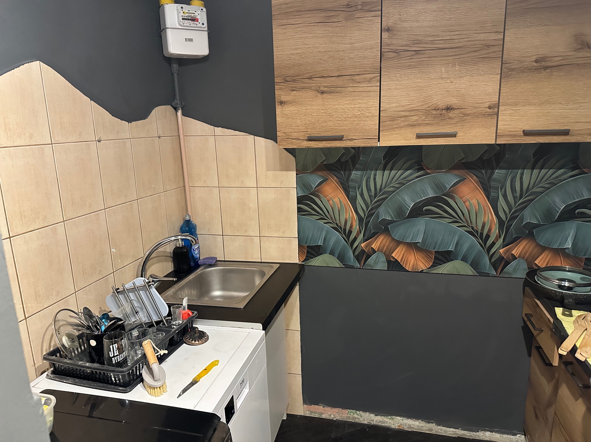 Fragment kuchni po odświeżeniu: beżowe płytki, ciemnoszara ściana, szafki z drewnianym wzorem i tapeta w liście. Widoczny zlew, suszarka na naczynia i licznik gazu.