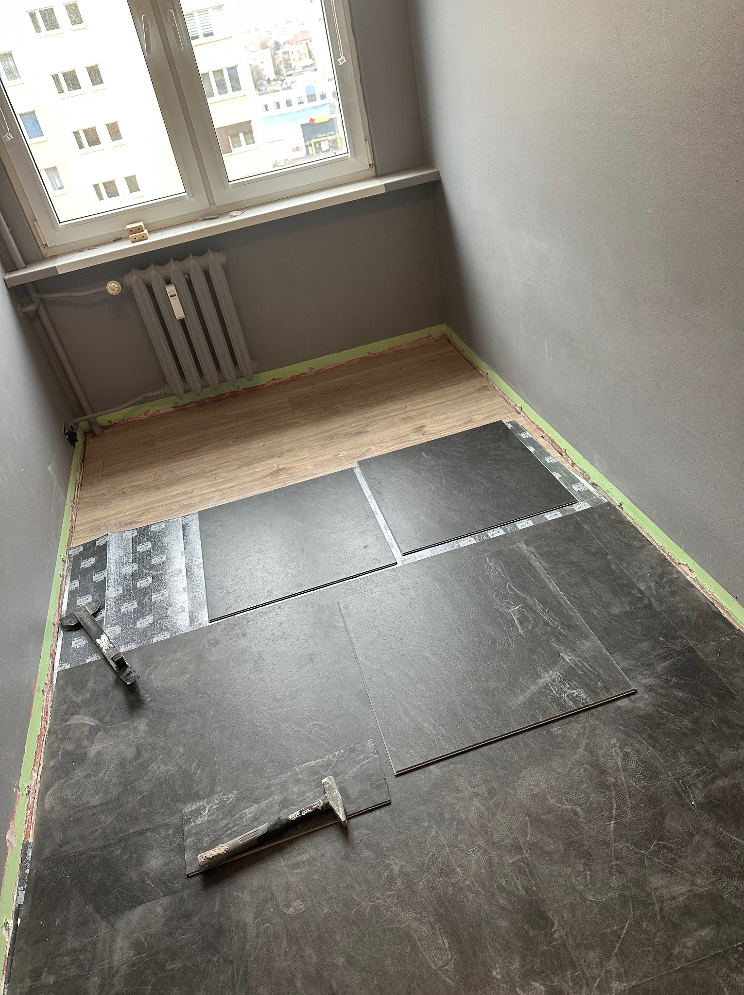 Ciemne płytki podłogowe układane w pokoju z szarą ścianą, widoczne narzędzia glazurnicze i fragment starej podłogi. Okno w tle.