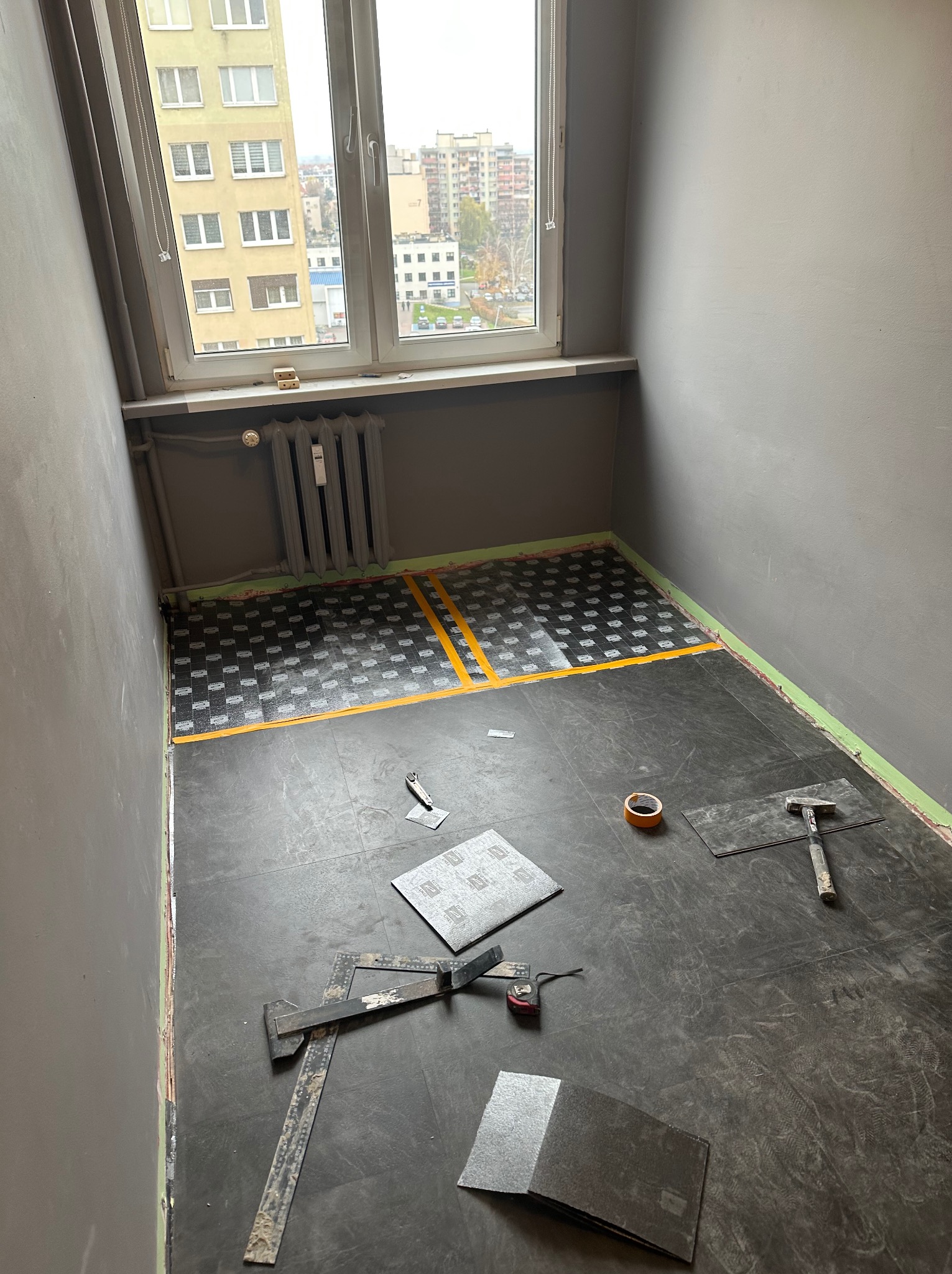 Częściowo ułożona podłoga z ciemnych paneli w pokoju, widoczne narzędzia glazurnicze i taśma malarska. Przy oknie grzejnik i widok na miasto.