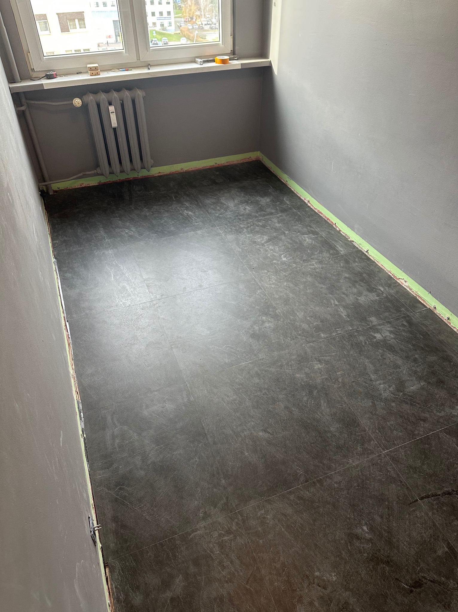 Szare płytki podłogowe ułożone w pustym pokoju z szarymi ścianami. Widoczny grzejnik i okno. Zabezpieczone ściany taśmą malarską. Ujęcie z góry, ukośne.