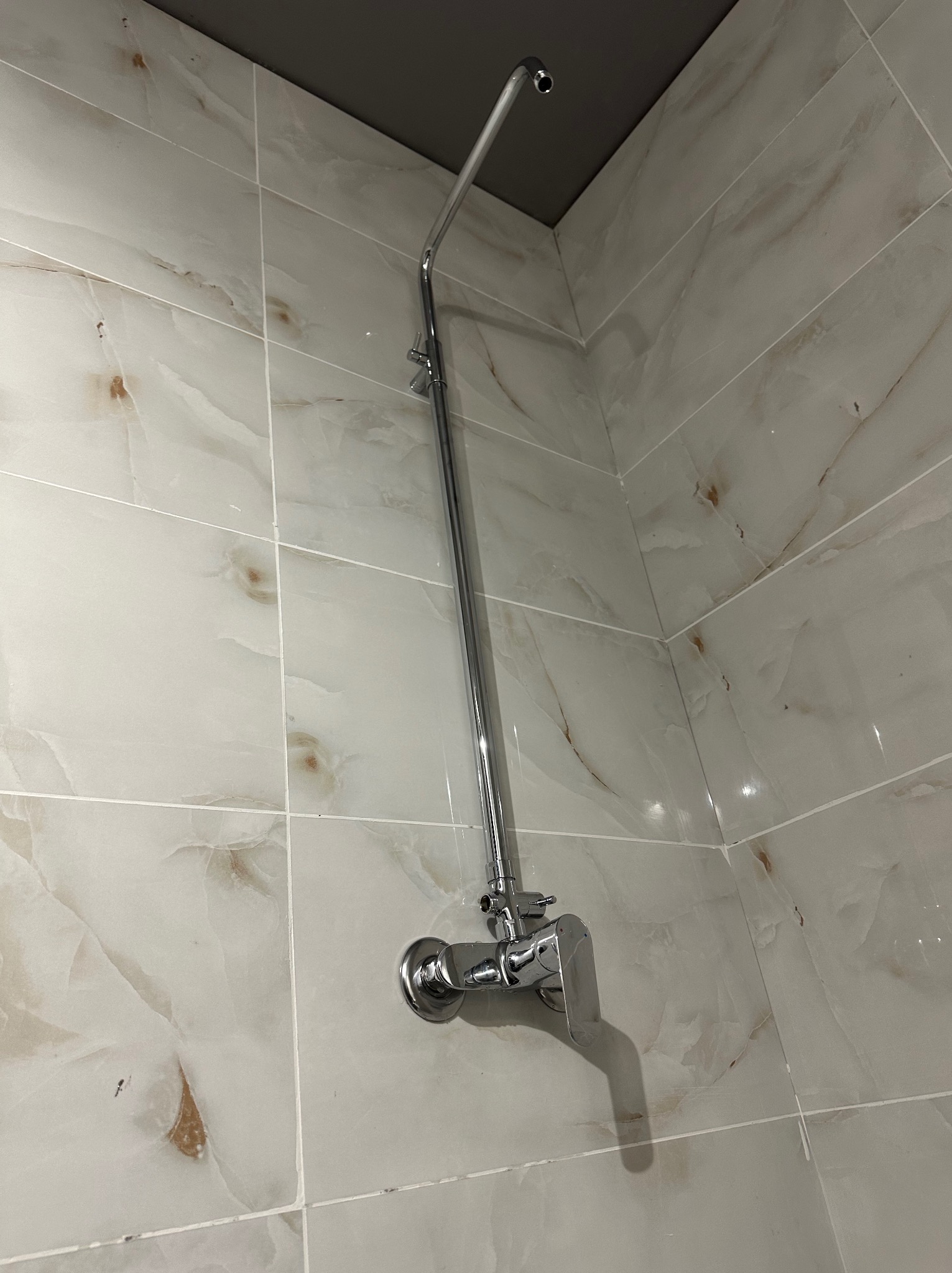 Błyszcząca, chromowana bateria natryskowa zamontowana na ścianie wyłożonej płytkami imitującymi marmur w odcieniach bieli i beżu. Widok z dołu, fragment sufitu.