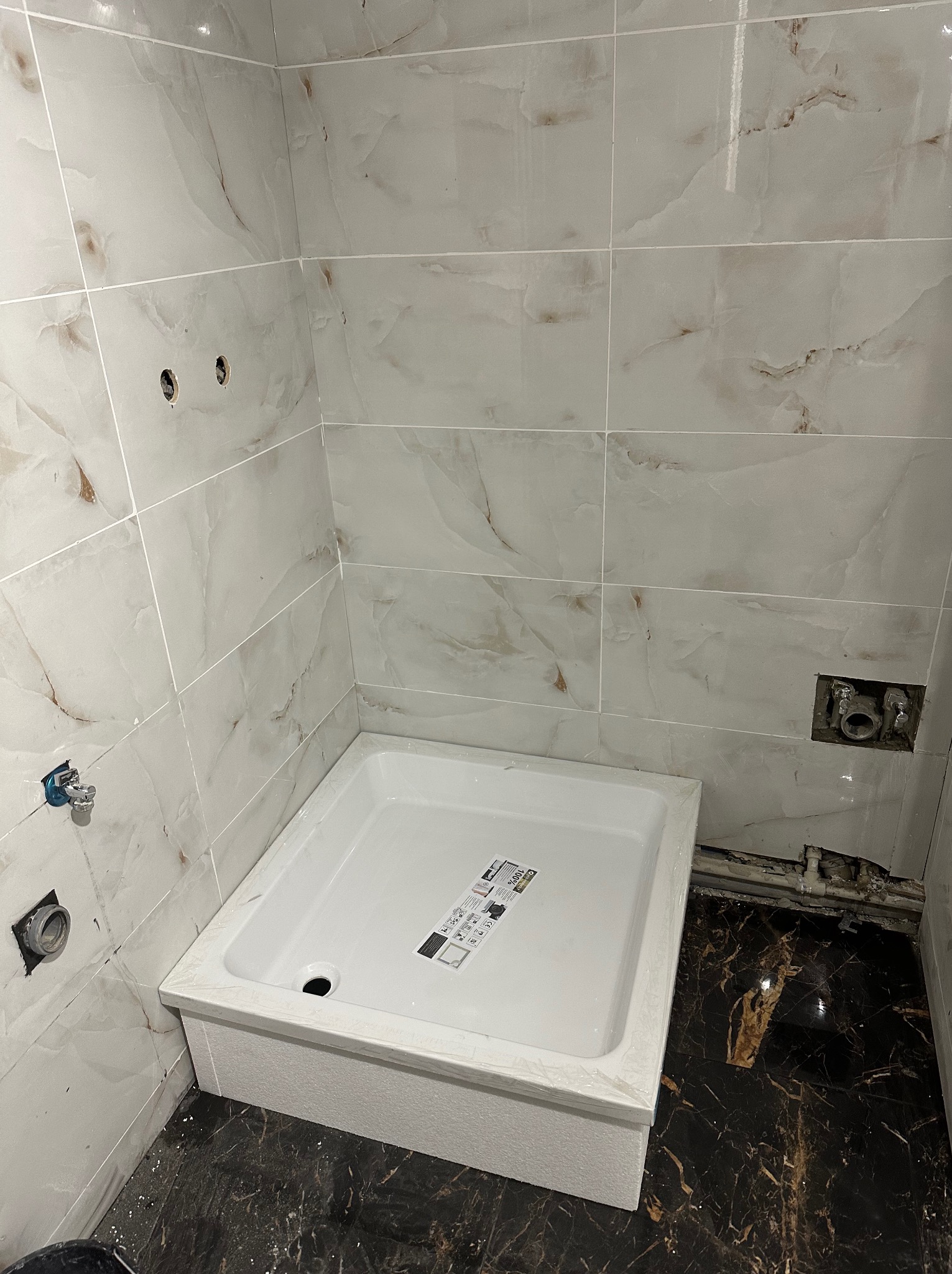 Biały brodzik w trakcie montażu w łazience z marmurowymi płytkami. Widoczne otwory na armaturę i instalacje. Podłoga wyłożona ciemnym marmurem.