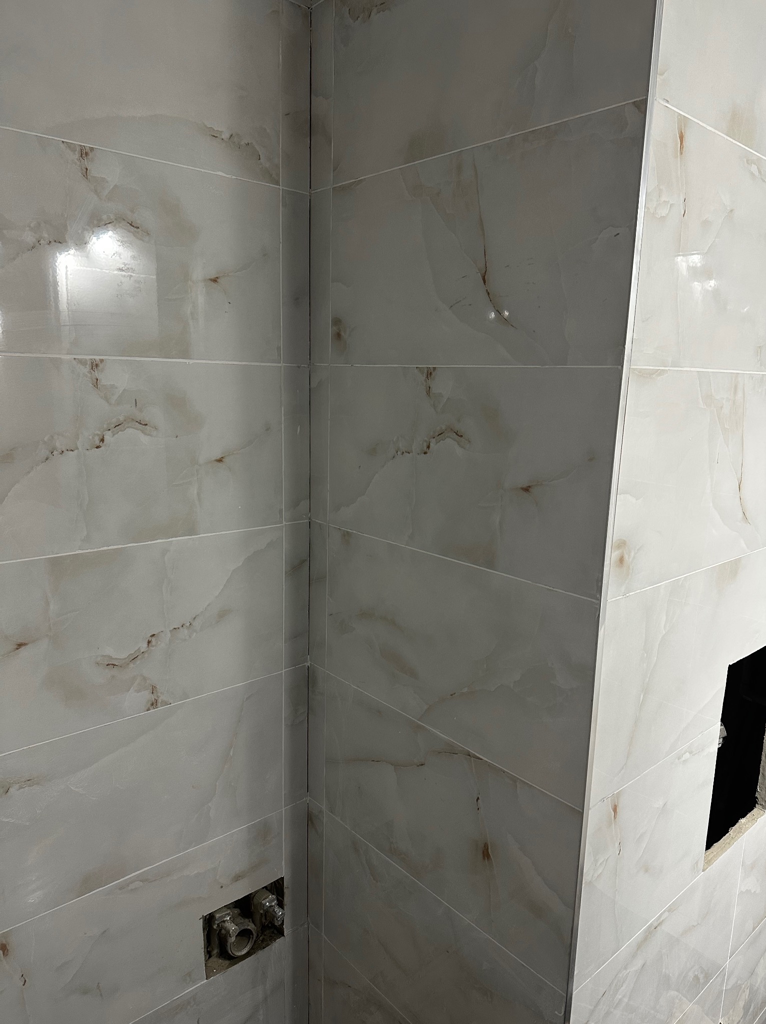 Wykończona ściana w łazience z jasnych, marmurkowych płytek. Widoczne fugi i instalacja wodna. Minimalistyczny design, gładka powierzchnia płytek.