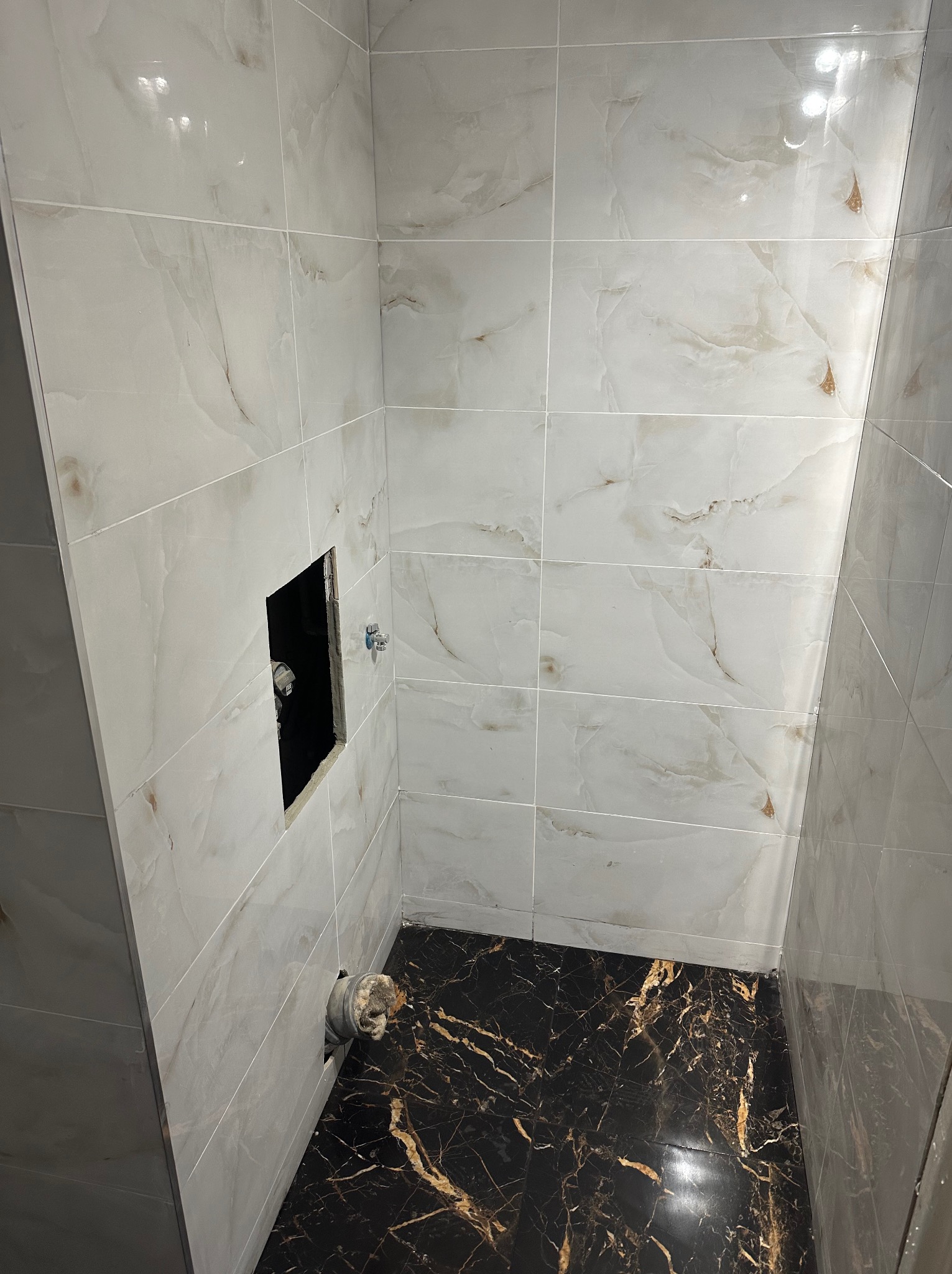 Wykończona kabina prysznicowa z marmurowymi płytkami na ścianach i podłodze. Widoczny otwór na baterię i odpływ. Nowoczesny design z kontrastującymi kolorami.