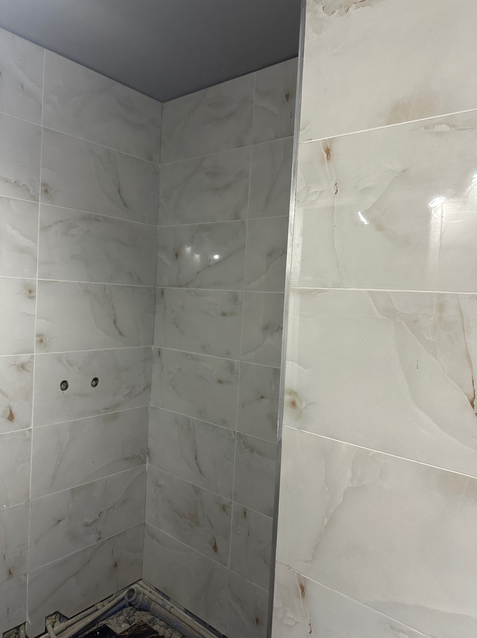 Wykończona ściana w łazience z jasnymi, marmurkowymi płytkami. Widoczne rury w dolnej części kadru i szara górna część ściany. Ujęcie z bliska.