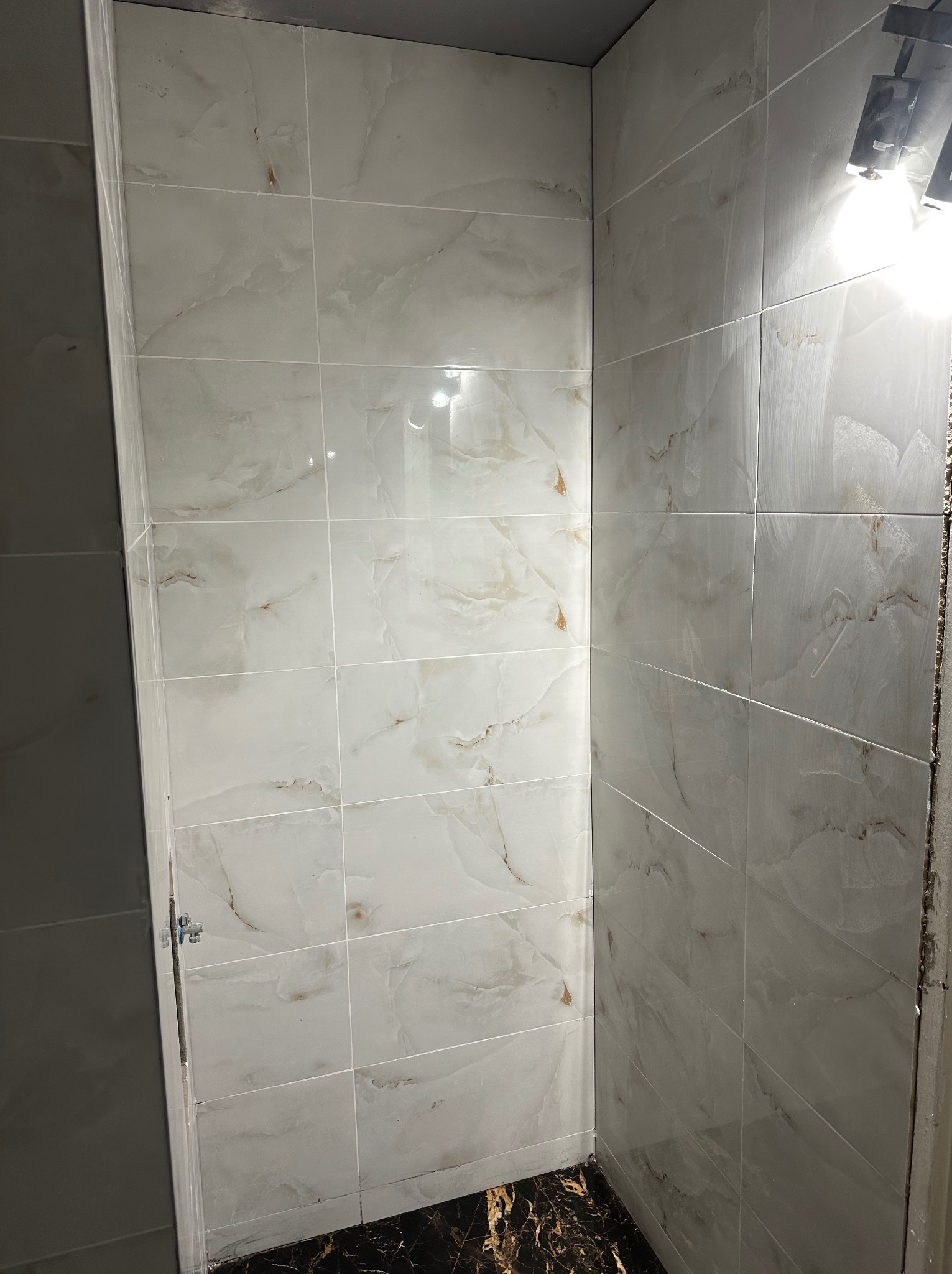 Wykończona ściana w łazience z jasnymi, marmurkowymi płytkami. Widoczny fragment podłogi z ciemnymi płytkami. Oświetlenie punktowe na górze kadru.