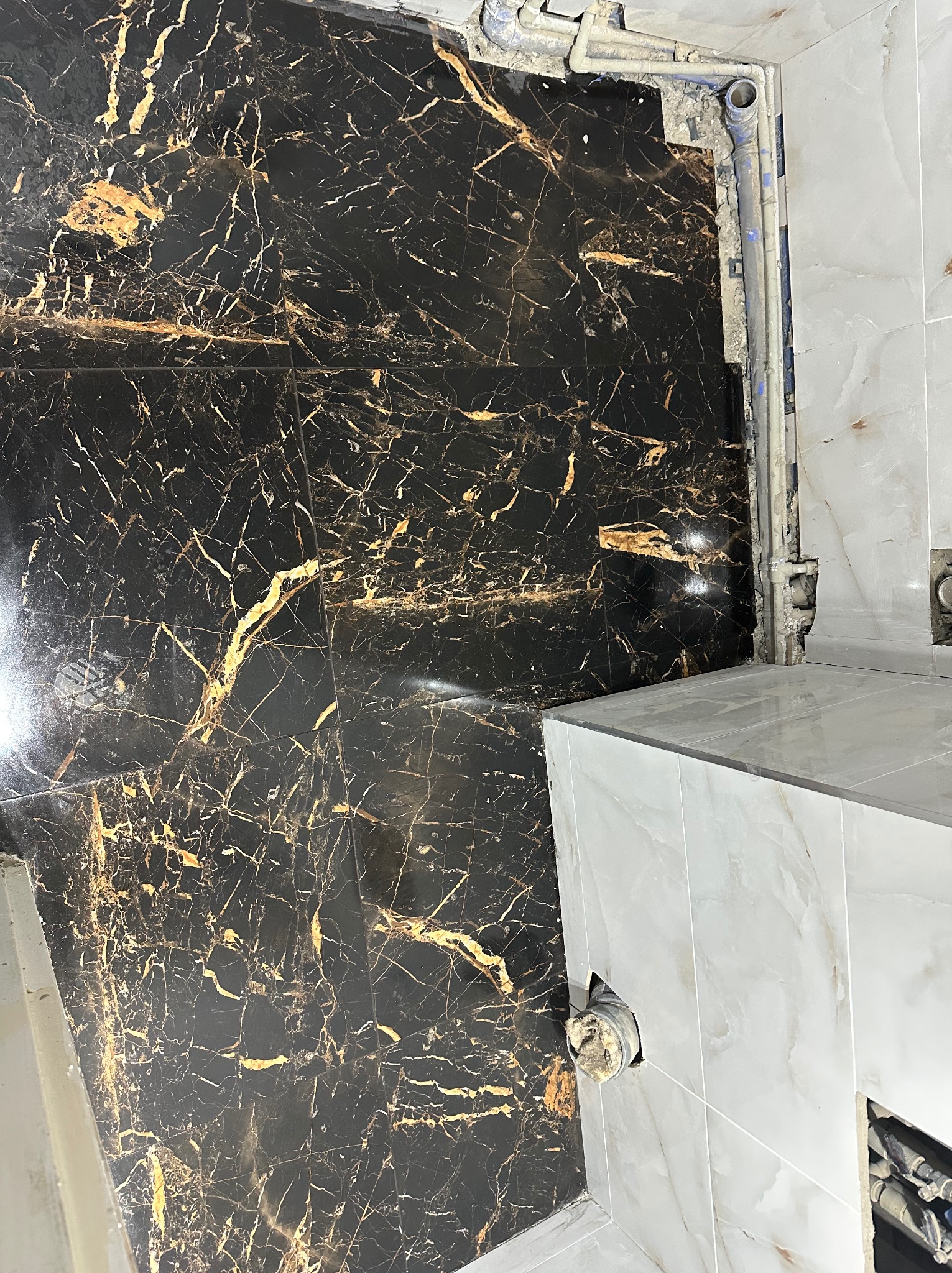 Ukończona podłoga z czarnych płytek imitujących marmur z złotymi żyłami, kontrastująca z jasnymi płytkami na ścianie. Widoczne rury instalacyjne.