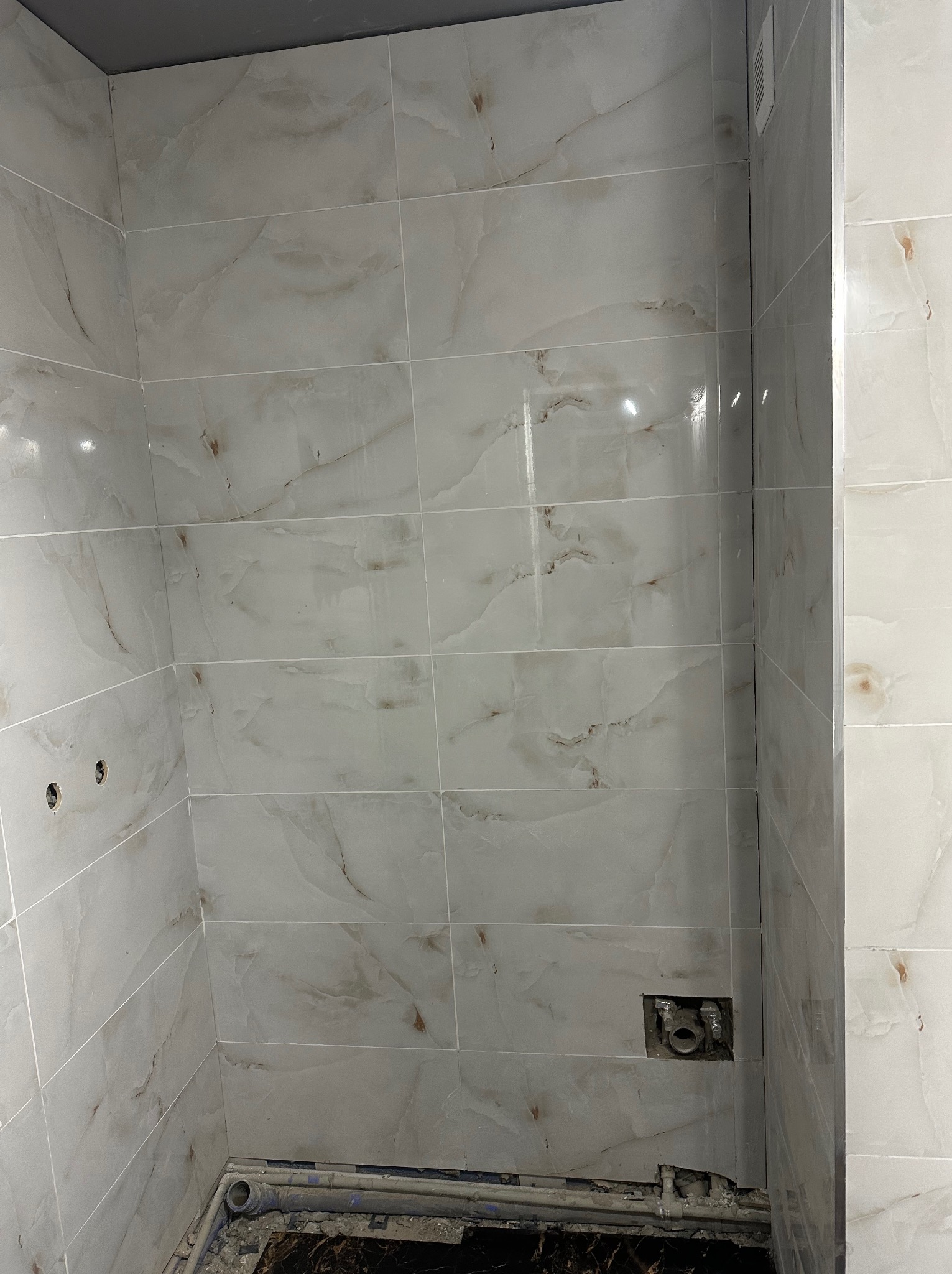 Wykończona ściana w łazience z jasnymi, marmurkowymi płytkami. Widoczne otwory na armaturę i fragment podłogi z instalacją. Minimalistyczny design.