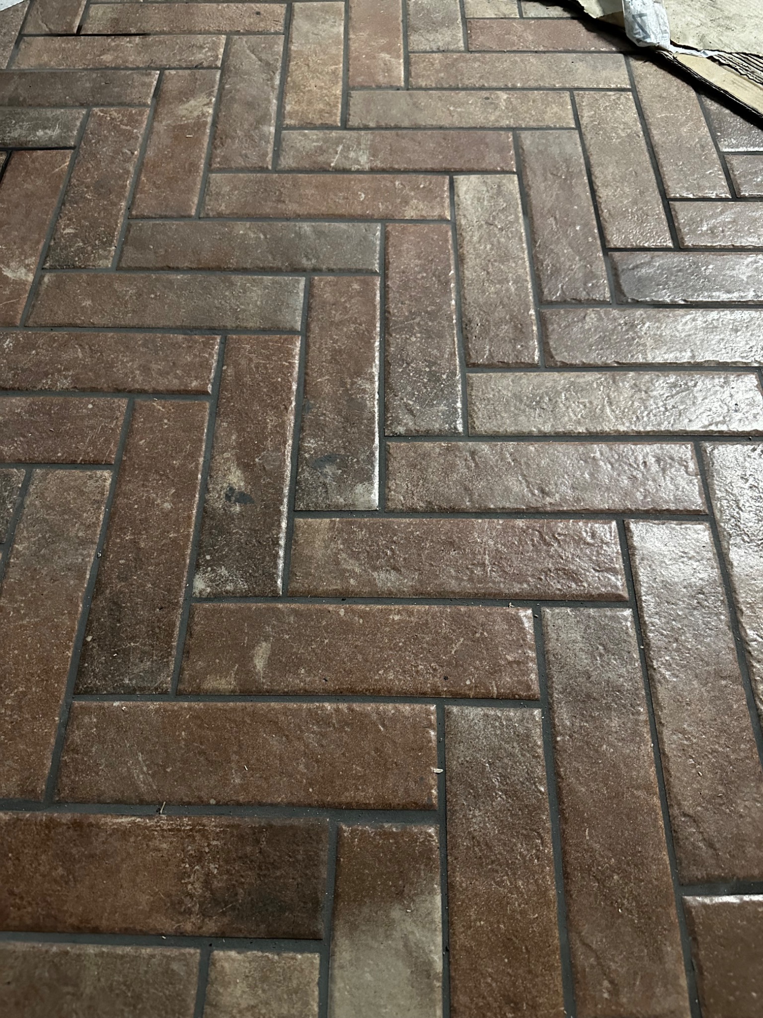 Ułożona w jodełkę, brązowa, ceramiczna podłoga z fugami w odcieniu grafitu. Widoczne drobne niedoskonałości i refleksy światła na powierzchni płytek.