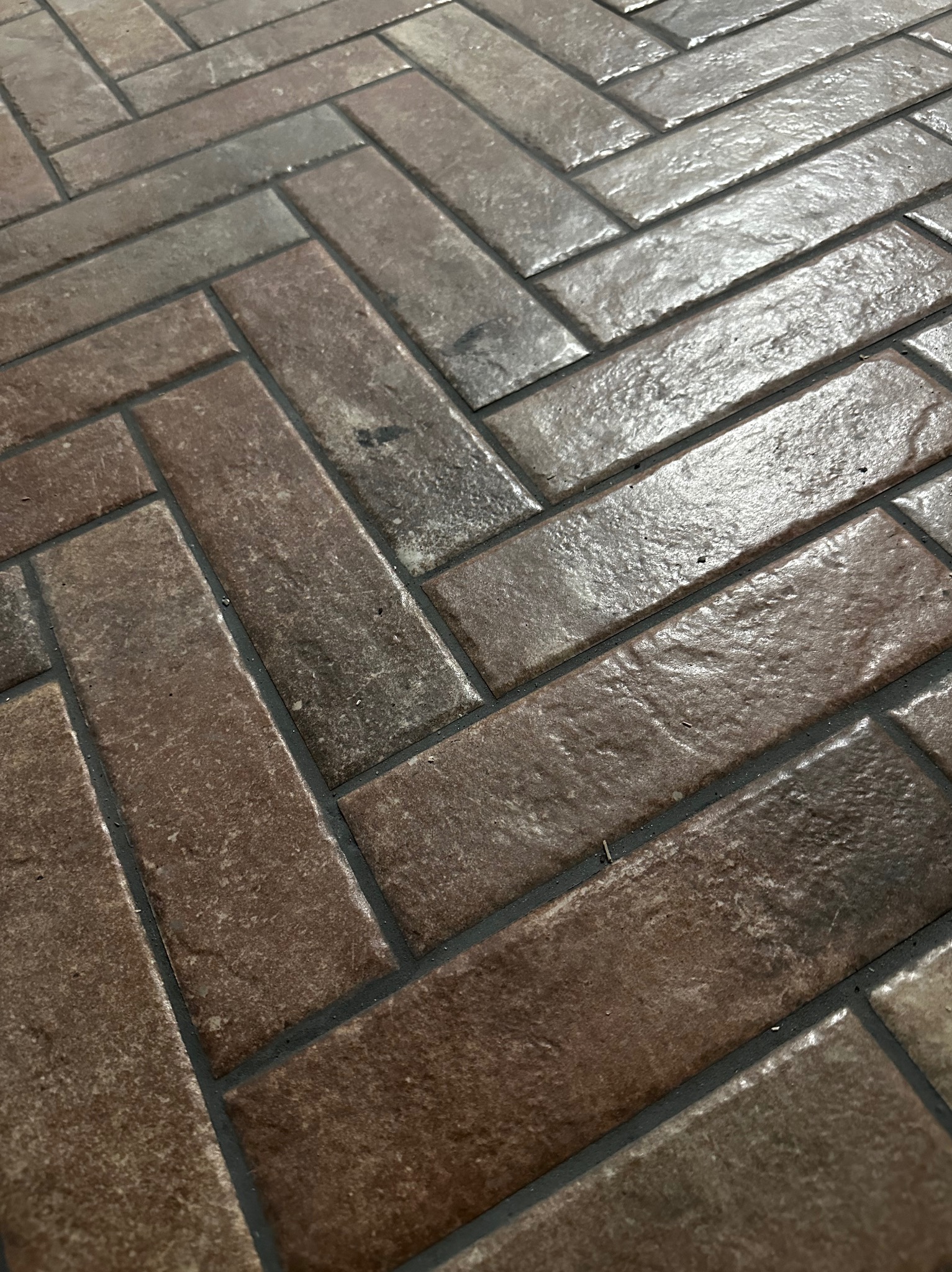 Ułożona w jodełkę, brązowo-szara, ceramiczna podłoga z fugą w odcieniu grafitu. Widok z góry, z bliska, ukazujący fakturę płytek i precyzję ułożenia.