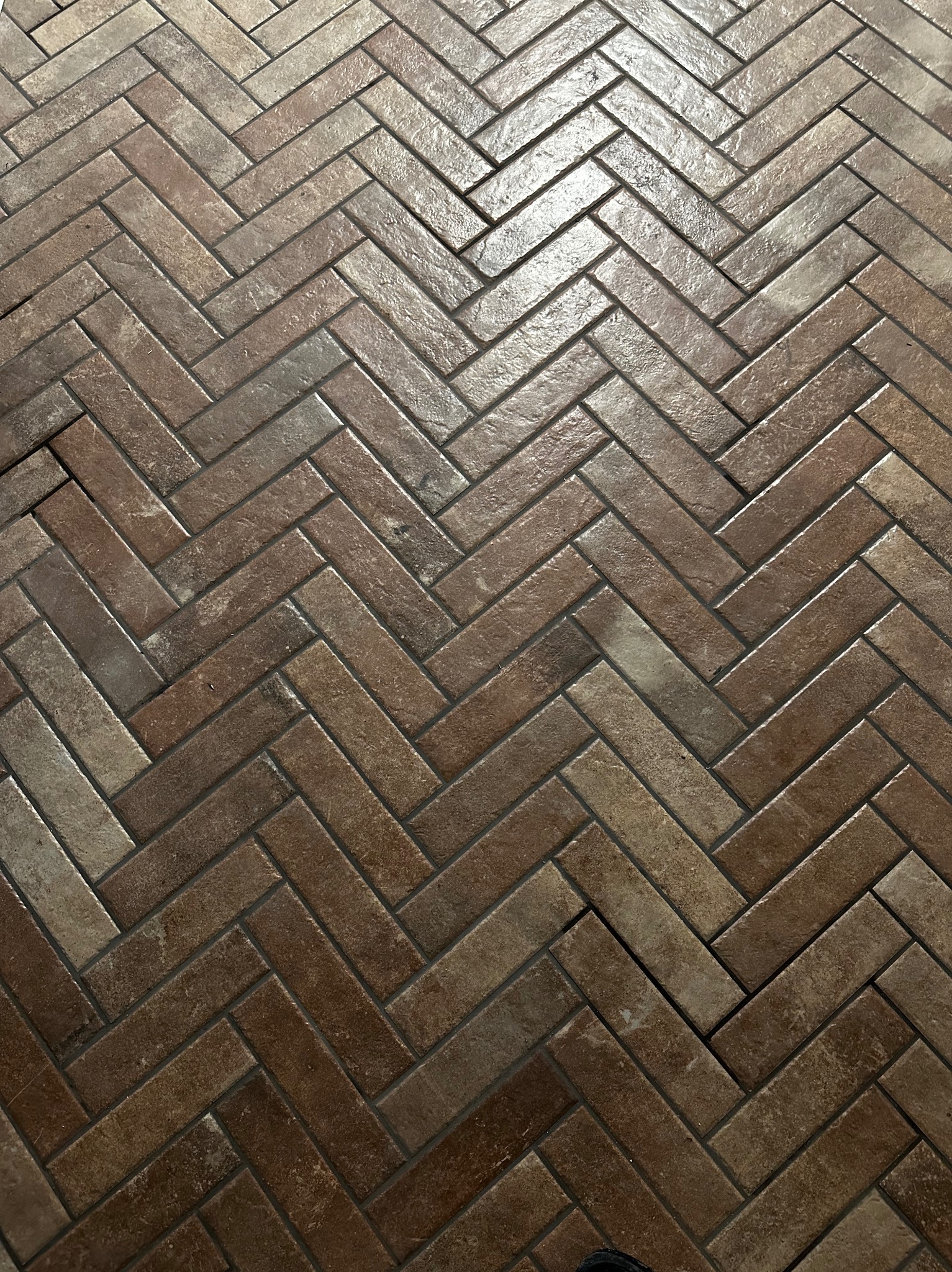 Ułożona w jodełkę, brązowa terakota podłogowa z delikatnym połyskiem, wypełniona ciemną fugą. Widoczny fragment buta w dolnym rogu kadru.