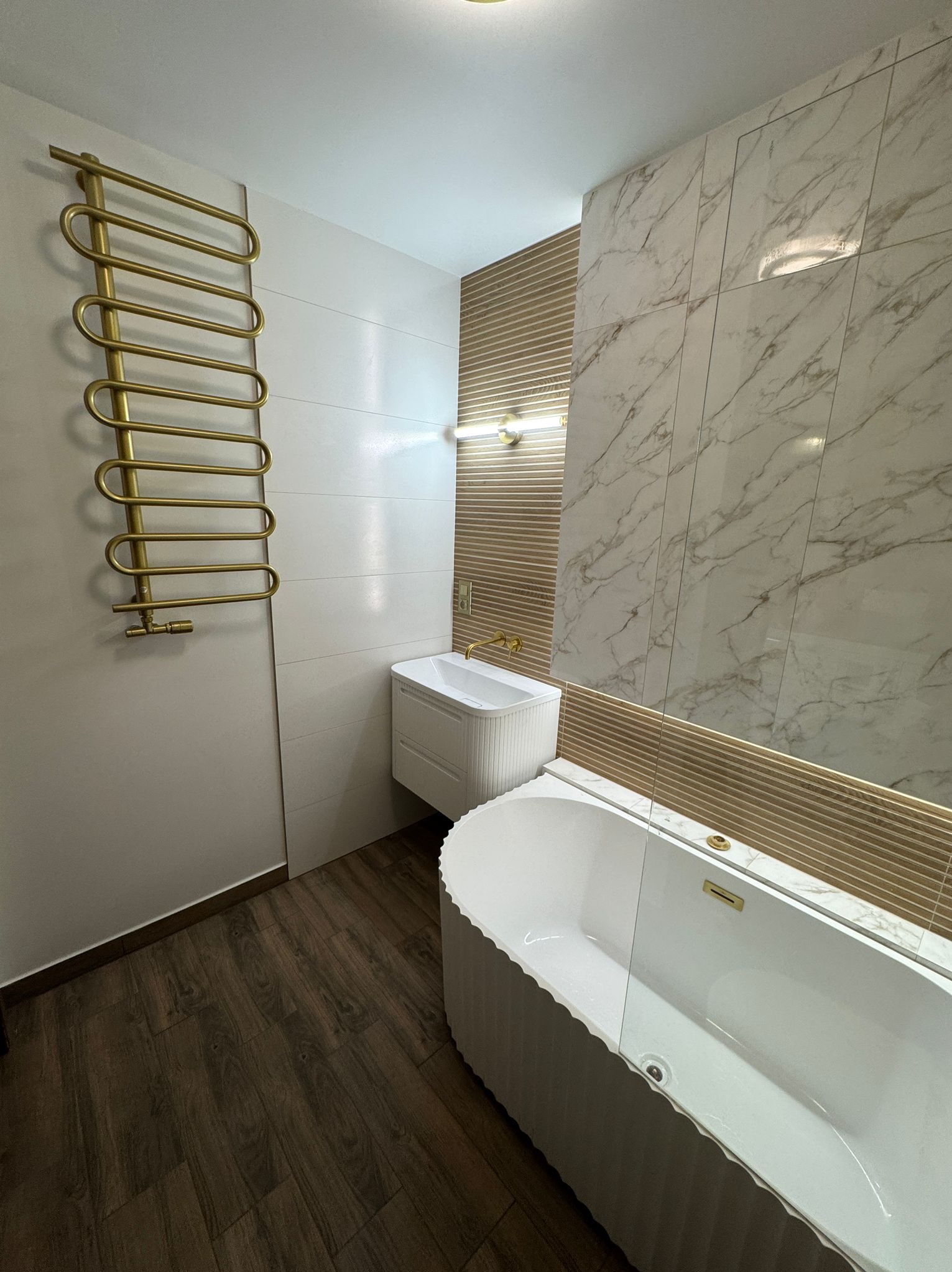 Nowoczesna łazienka w Warszawie: biała wanna z dekoracyjną obudową, umywalka z szafką, złoty grzejnik drabinkowy i płytki imitujące marmur.
