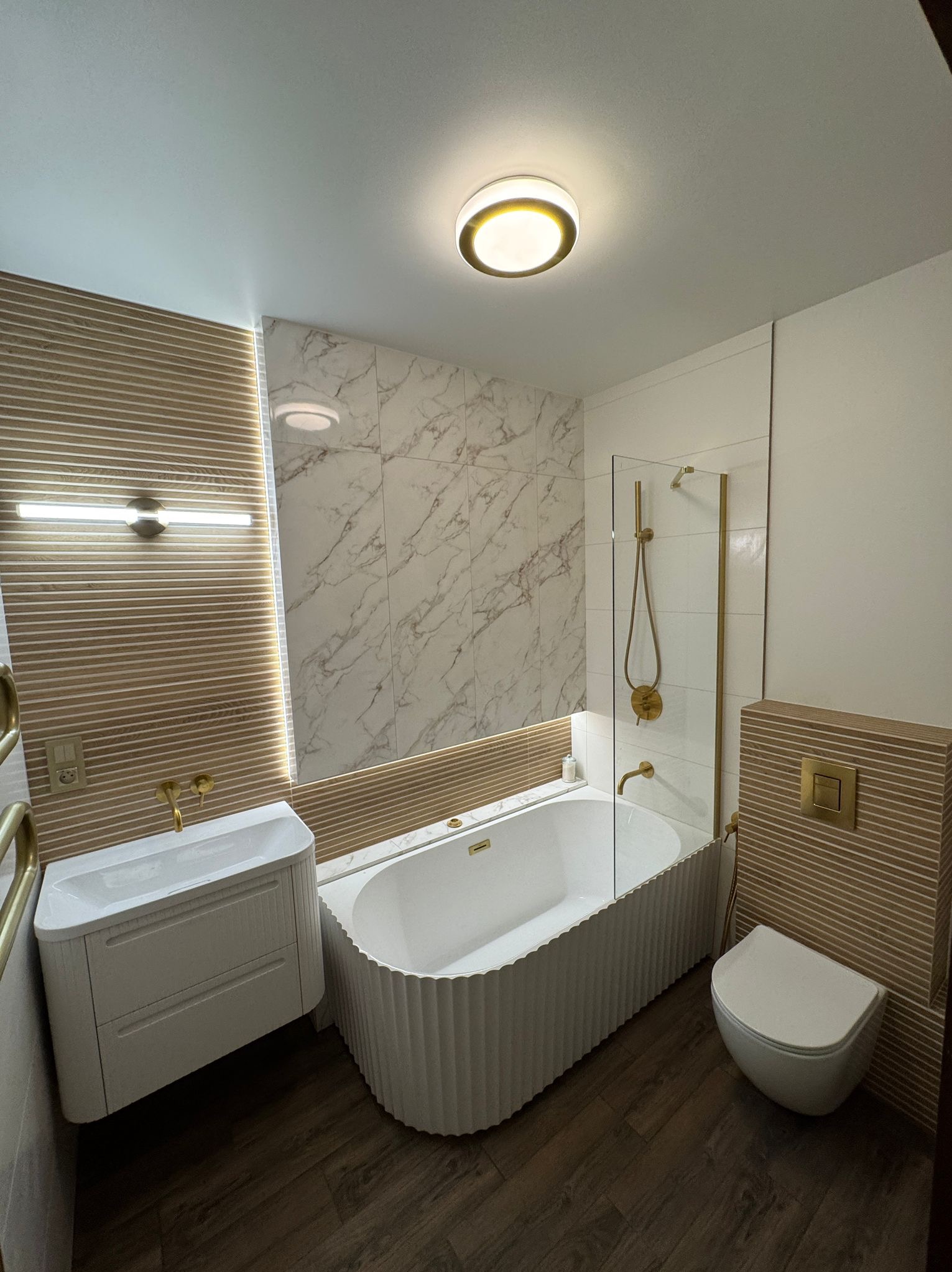 Nowoczesna łazienka z wanną o falistym wykończeniu, marmurowymi płytkami i złotą armaturą. Umywalka z szafką i podwieszana toaleta tworzą spójną całość.