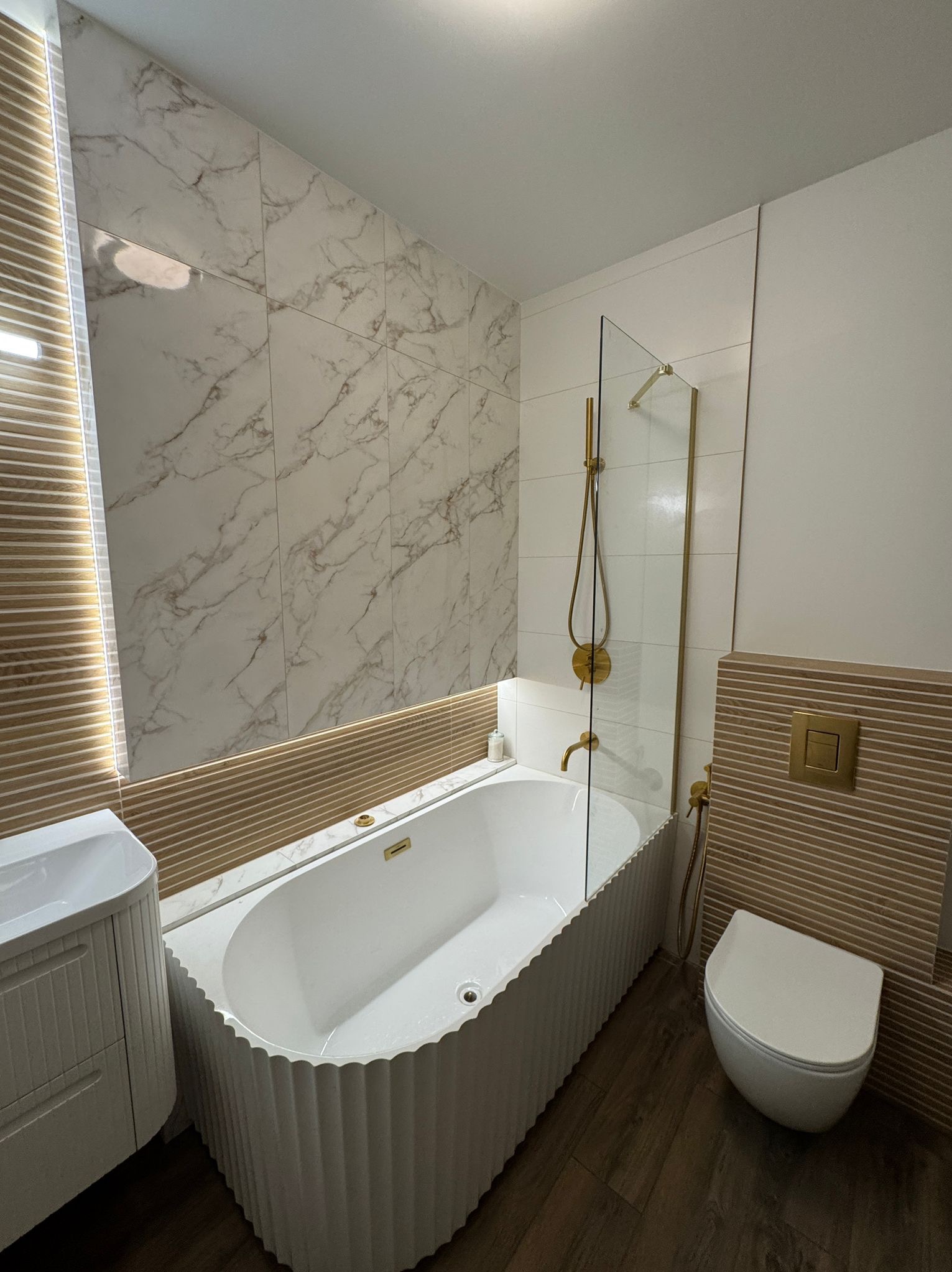 Elegancka łazienka z wanną o falistej obudowie, marmurowymi płytkami i złotymi akcentami. Kabina prysznicowa ze szkła hartowanego. Nowoczesny design.