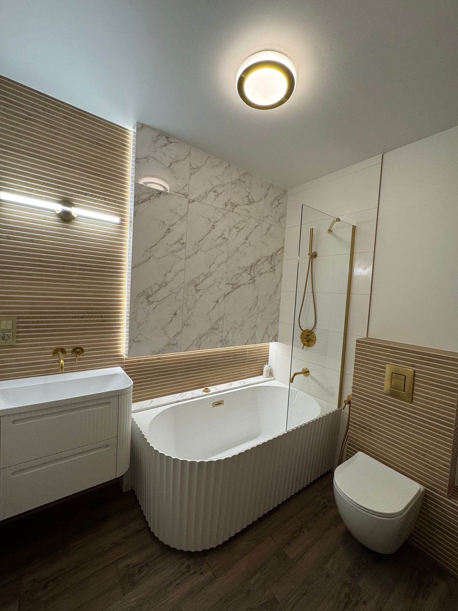 Nowoczesna łazienka z wanną, umywalką i toaletą w odcieniach bieli, beżu i złota. Uwagę zwracają ryflowane fronty mebli i marmurowe płytki.