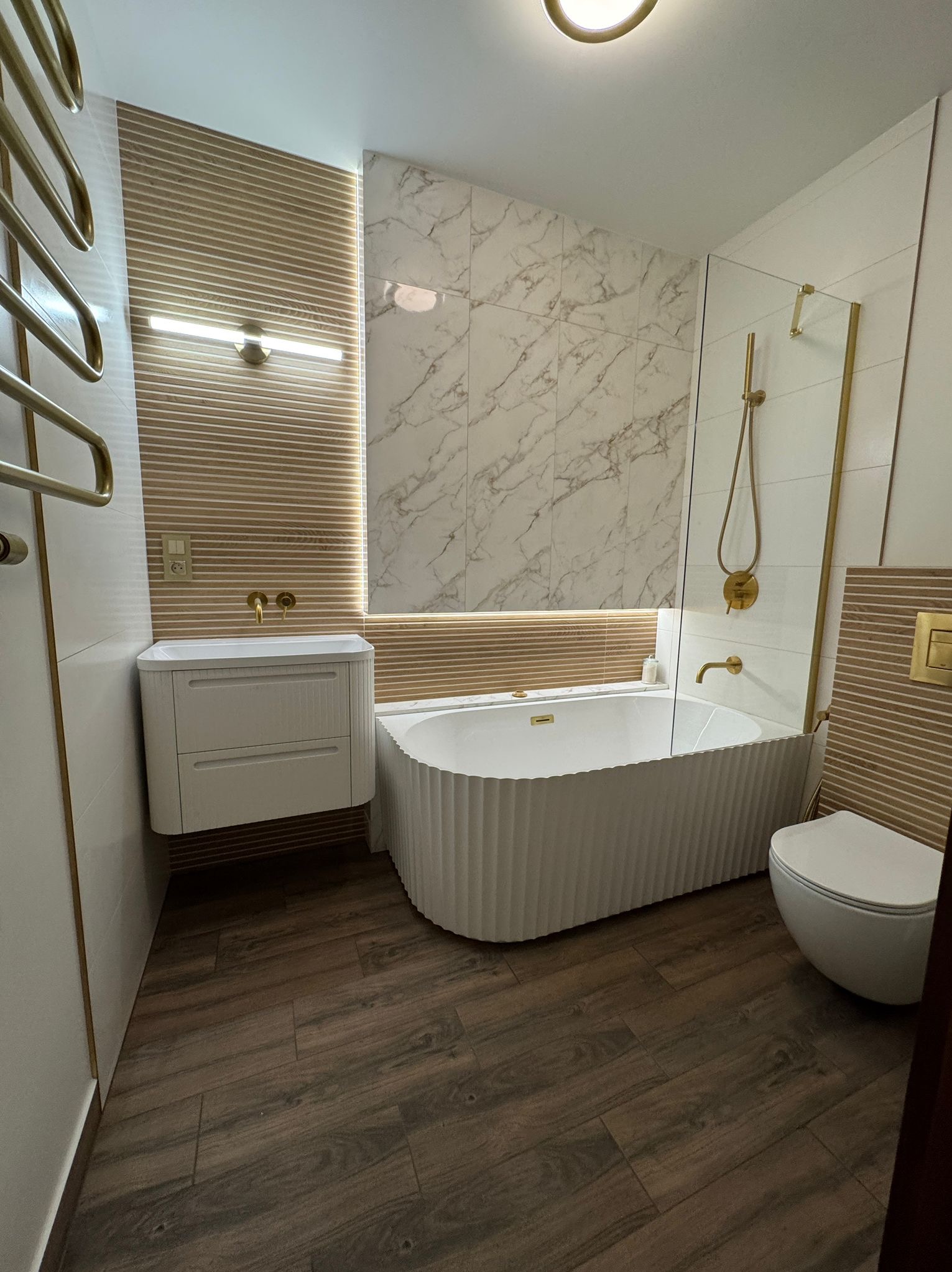 Nowoczesna łazienka z wanną o falistej obudowie, marmurowymi płytkami i złotymi akcentami. Umywalka z szafką w kolorze białym, drewnopodobna podłoga i grzejnik drabinkowy.
