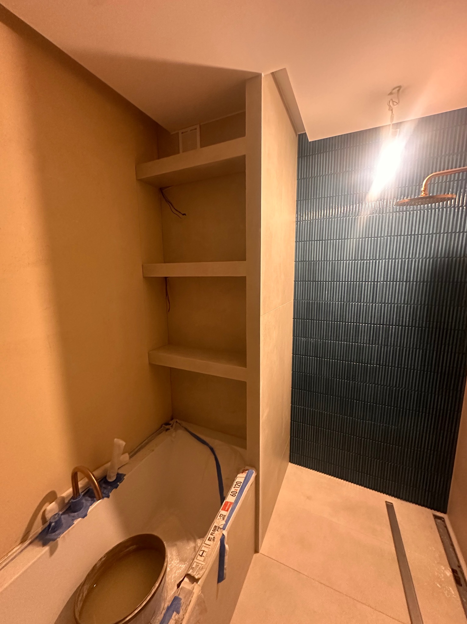 Remont łazienki w trakcie: zabudowa z półkami, ułożone płytki na ścianie, przygotowana wanna z baterią. Widoczne materiały budowlane i narzędzia.