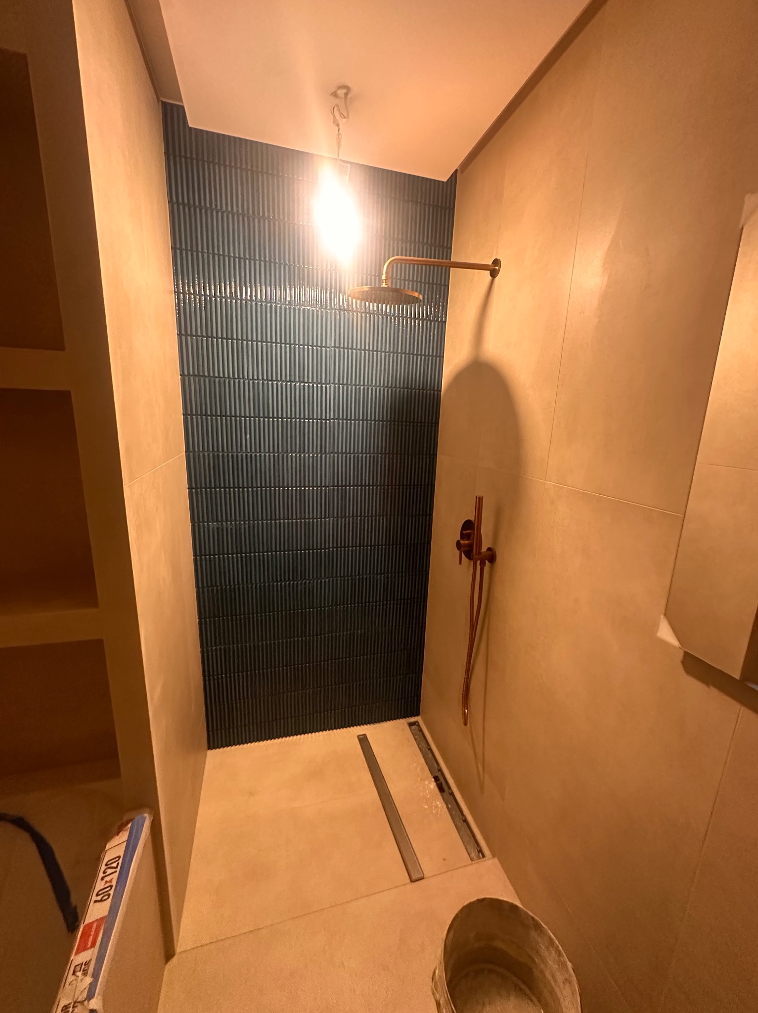 Nowoczesna łazienka w trakcie wykańczania: pionowe, ciemne płytki na ścianie, miedziana armatura, odpływ liniowy w podłodze. Widoczne materiały budowlane.