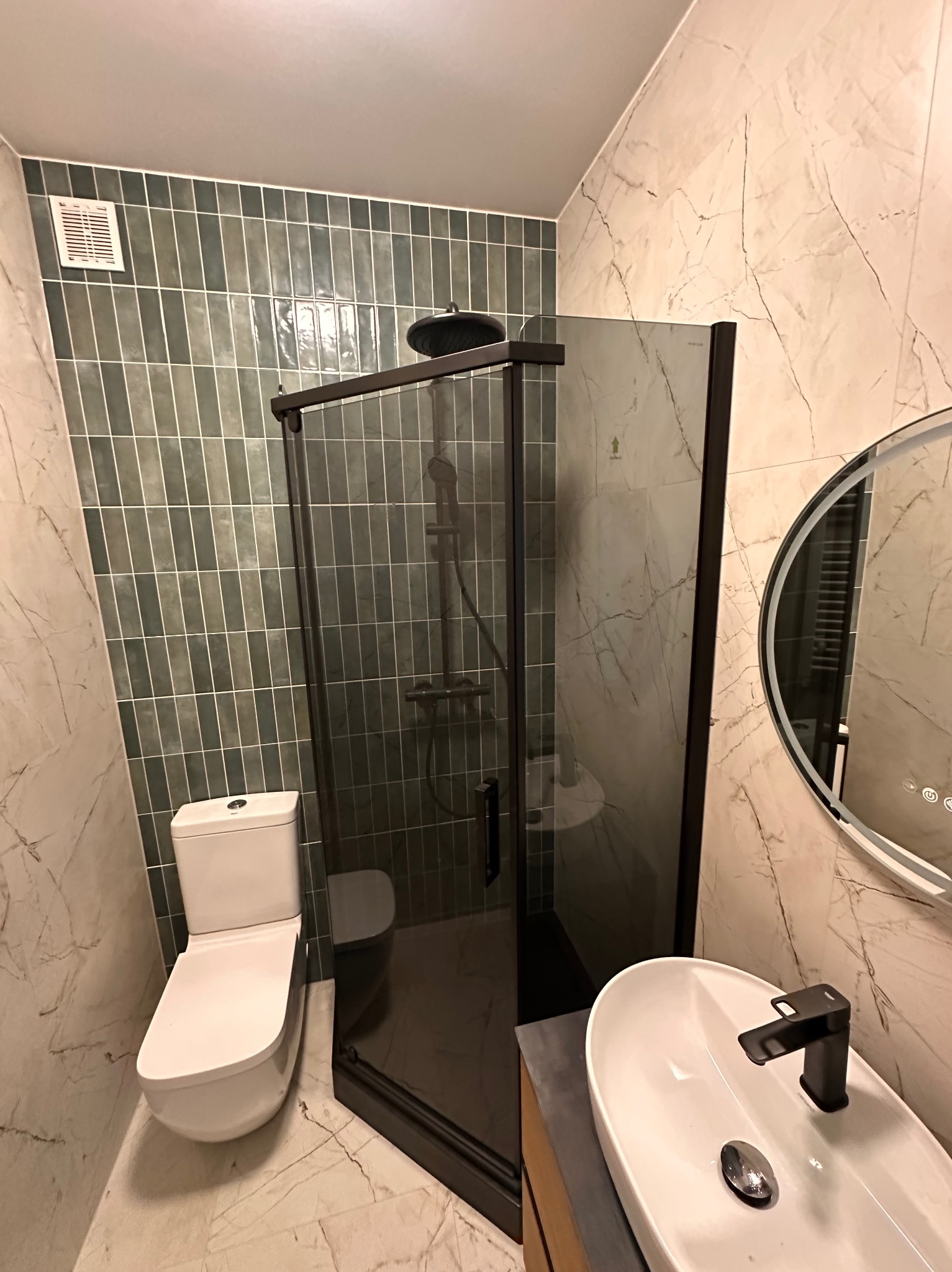 Nowoczesna łazienka z kabiną prysznicową typu walk-in, umywalką nablatową i toaletą. Ściany wyłożone płytkami imitującymi marmur i zielonymi płytkami w układzie pionowym.