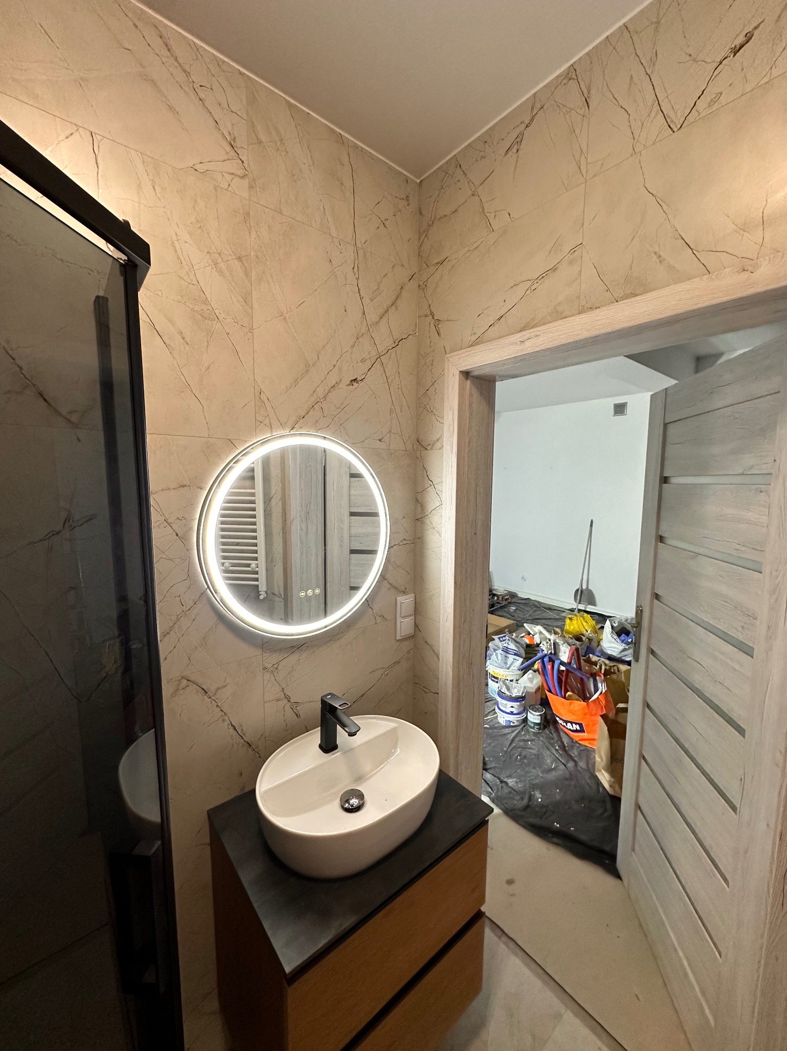 Wykończona łazienka z umywalką na szafce i okrągłym lustrem z podświetleniem LED. W tle widoczne materiały budowlane, sugerujące trwający remont.