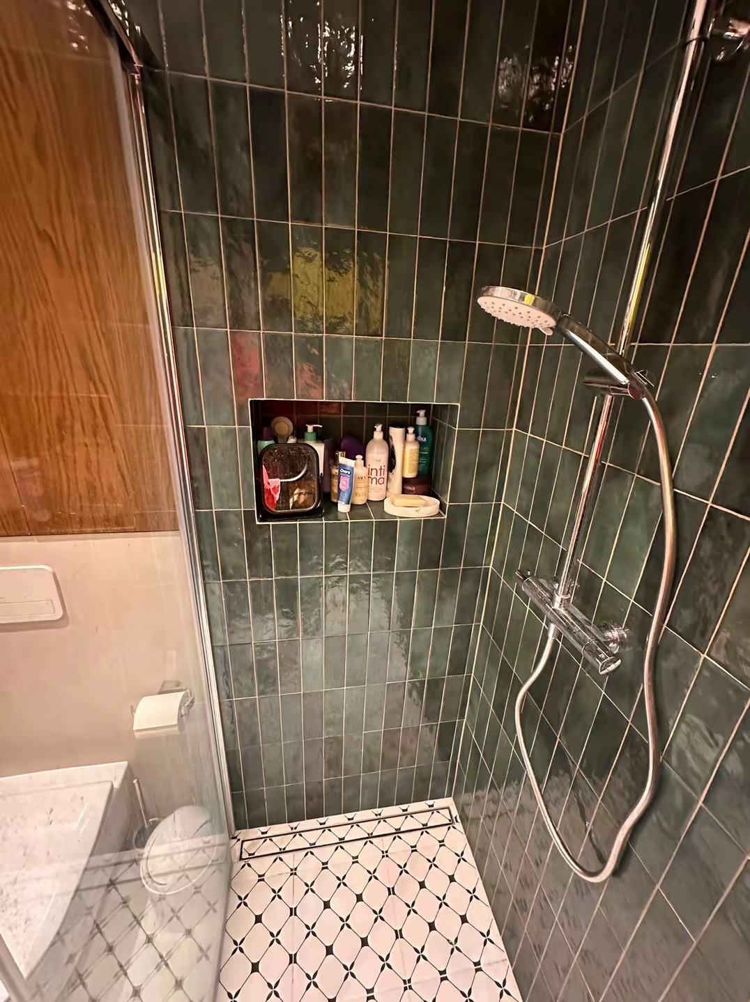 Wykończona kabina prysznicowa z zielonymi, pionowo ułożonymi płytkami. Wnęka z kosmetykami i biało-czarna mozaika na podłodze. Nowoczesna armatura.