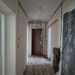 PS-ELECTRO - Korytarz w trakcie remontu z widocznymi przewodami elektrycznymi na ścianach i suficie. Częściowo zabezpieczone ściany folią malarską. Widoczne kable do oświetlenia.