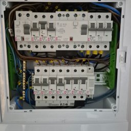 PS-ELECTRO - Rozdzielnica elektryczna w białej obudowie, widoczne bezpieczniki, przewody i złączki. Instalacja schludna, z oznaczeniami. Detal montażu wewnątrz budynku.