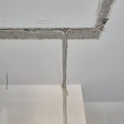 PS-ELECTRO - Białe kable elektryczne ułożone w bruzdach na suficie i ścianie w trakcie prac wykończeniowych. Widoczne przewody i puszki instalacyjne.